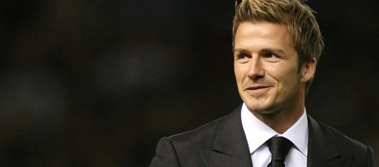 Ein Mann in einem schwarzen Anzug und weißem Hemd lächelt leicht vor dunklem Hintergrund.