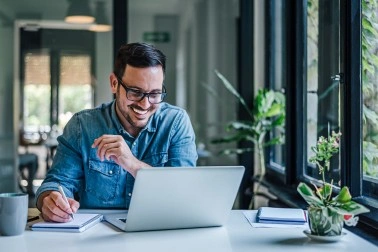 Smiling man in glasses and denim shirt writing notes while looking at laptop in bright room.