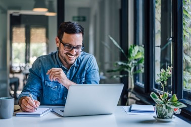 Smiling man in glasses and denim shirt writing notes while looking at laptop in bright room.