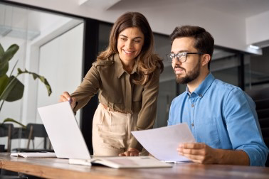 Woman shows laptop screen to man holding paper at a wooden table in modern office.