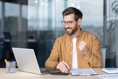 Smiling man in brown shirt celebrating while working on a laptop at a desk with documents.