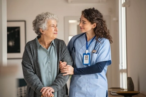 Smiling young female nurse or caregiver in blue scrubs with a stethoscope and ID badge offers support by holding the arm of an older woman with gray curly hair wearing a gray cardigan and light blue shirt, indoors in a warmly lit home setting.