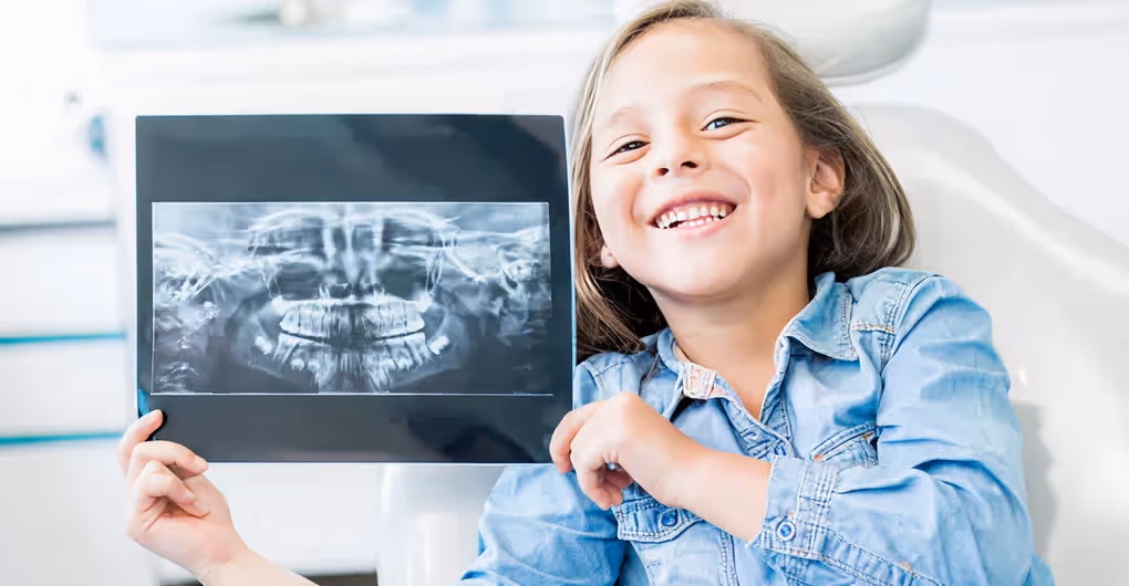 Smiling young girl holding up a dental panoramic X-ray showing her teeth and jaw structure.