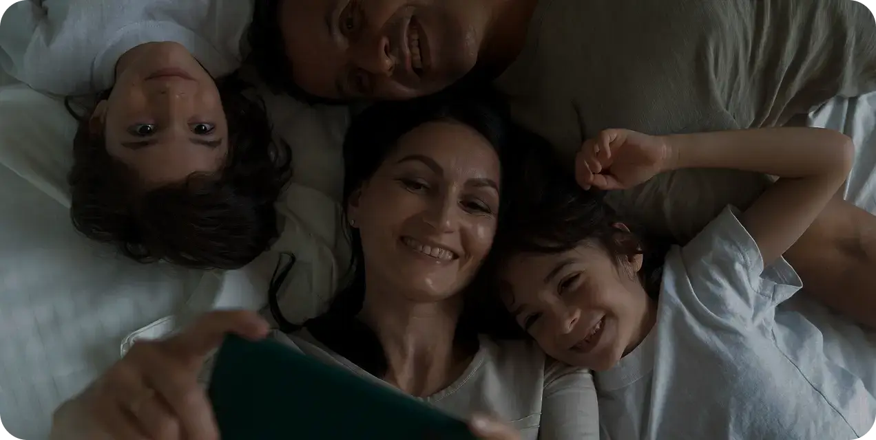 A happy family lying together on a bed, smiling while taking a selfie with a smartphone.