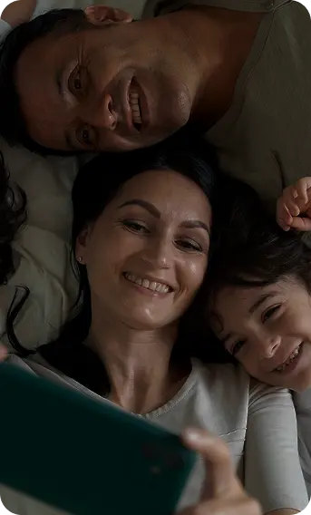 A happy family lying together on a bed, smiling while taking a selfie with a smartphone.