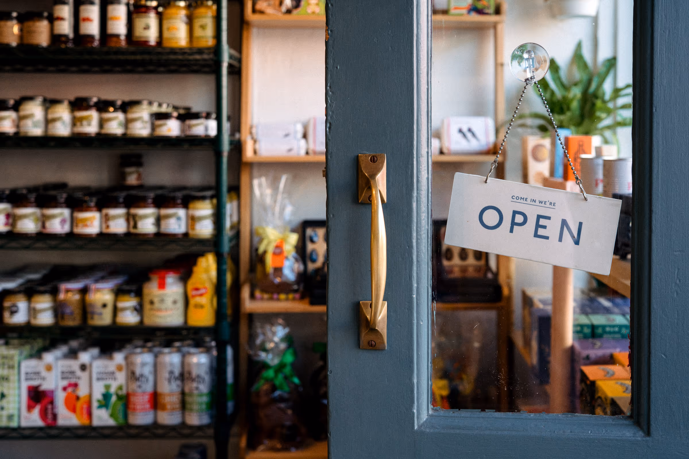 "OPEN" sign hangs on the open door of The Good Food Shop with deli produce on shelves in the background