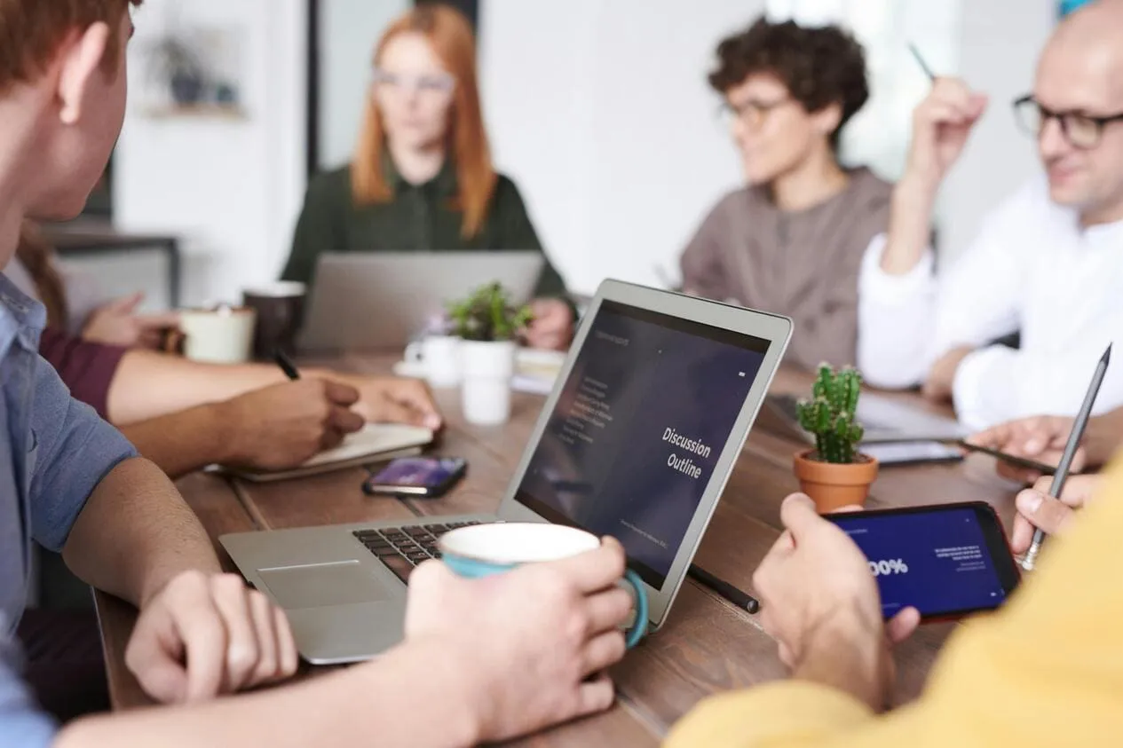 Mehrere Personen sitzen um einen Holztisch in einem modernen Büro, mit Laptops, Tablets und Notizbüchern.