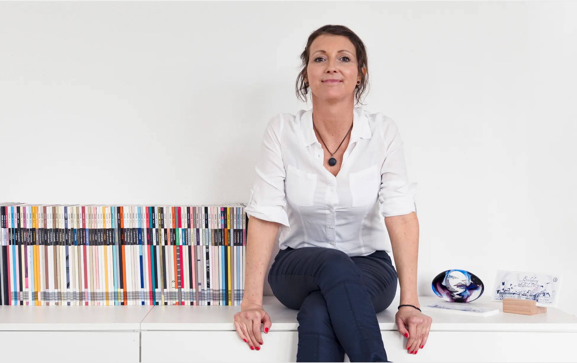Elisabeth Poth, in weissem Hemd und dunkler Hose, sitzt entspannt auf einem weissen Sideboard vor weissem Hintergrund. Links von ihr steht eine lange Reihe farblich sortierter Bücher, rechts dekorative Gegenstände wie ein Kristallkunstwerk und Karten. Sie blickt freundlich in die Kamera.