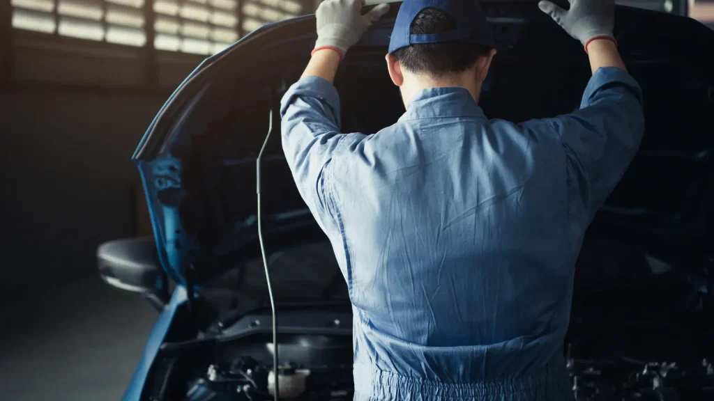 Mechanic in blue coveralls examining a car engine under the hood in a dimly lit garage