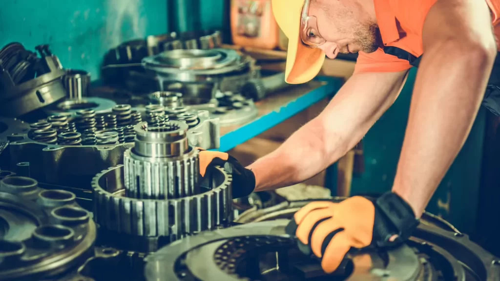Transmission repair shops featuring a mechanic in gloves and a hard hat working on a disassembled transmission system in a workshop