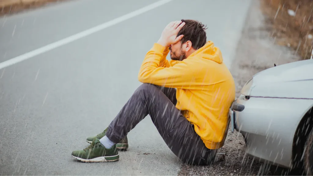 Transmission service needed as a frustrated man in a yellow hoodie sits beside his broken-down car in the rain.