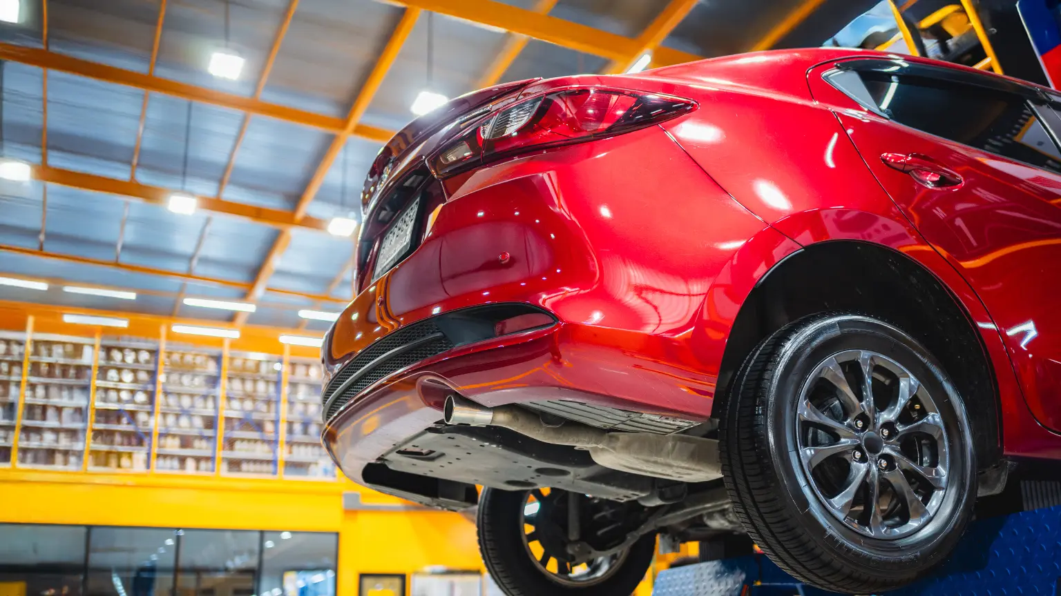 Car repair in progress with red vehicle lifted on hydraulic lift inside brightly lit auto shop