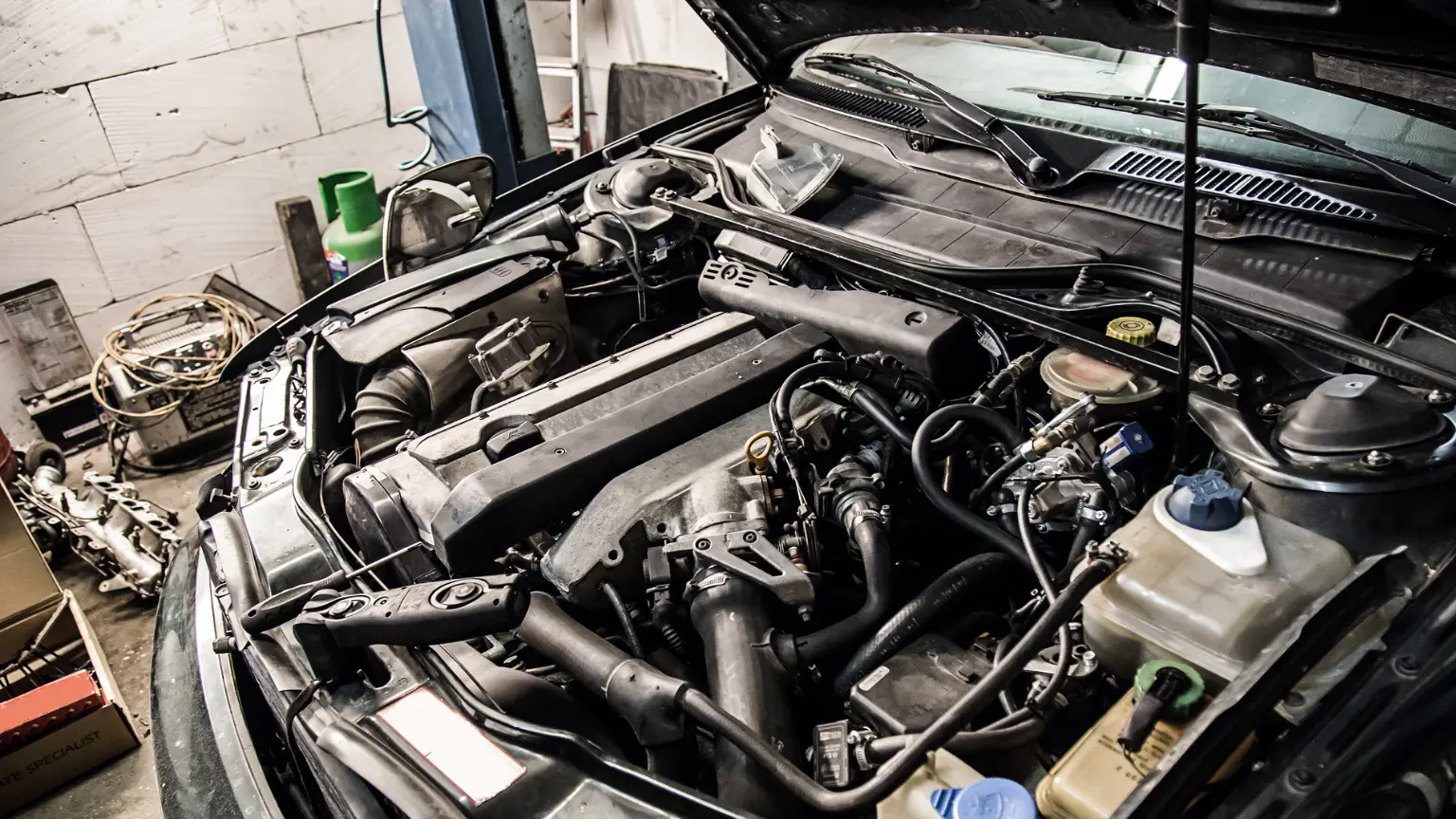 Car repair in progress with engine exposed inside a cluttered garage workspace