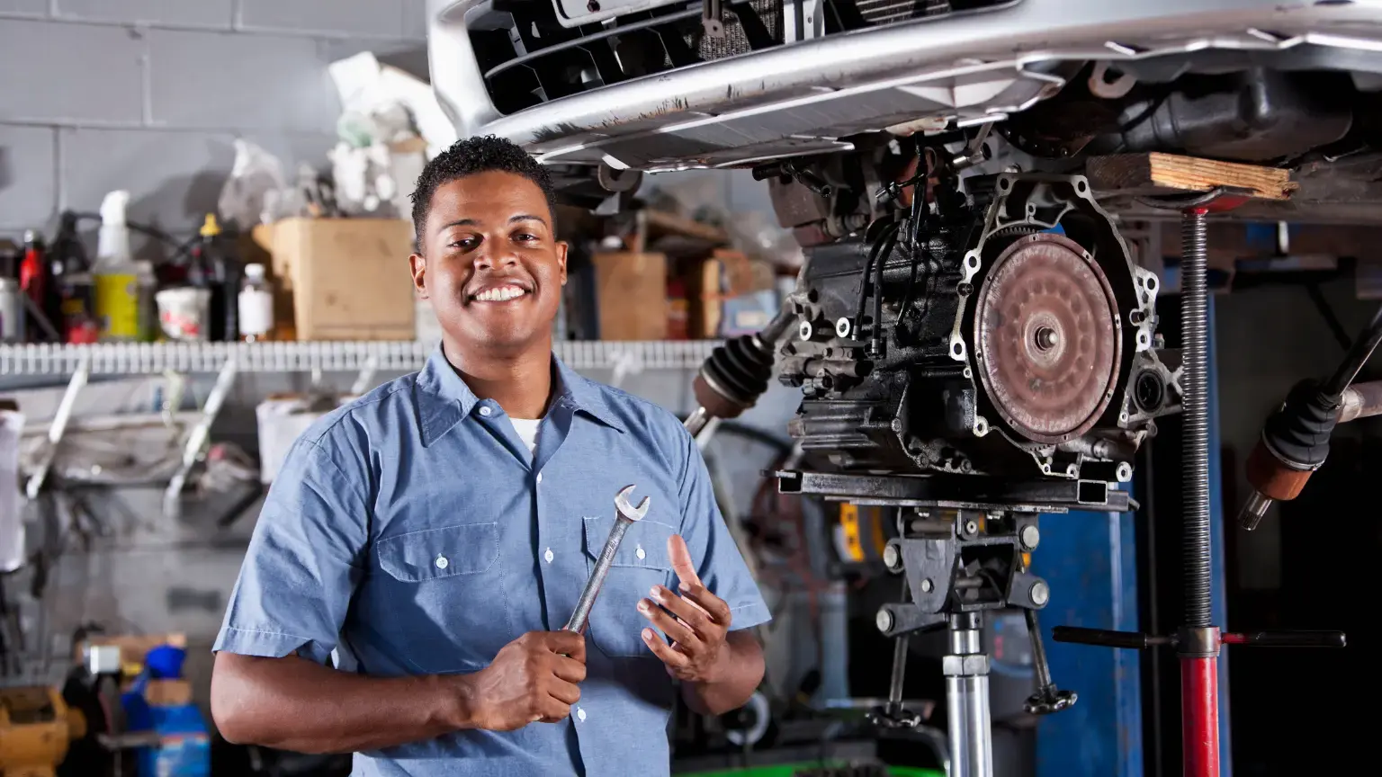 Transmission mechanic standing beside a car with exposed transmission system in an auto repair garage.