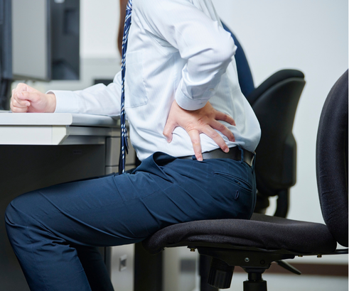 Un homme en chemise blanche et cravate assis sur une chaise de bureau, tenant son dos avec une main, semblant avoir mal au dos.