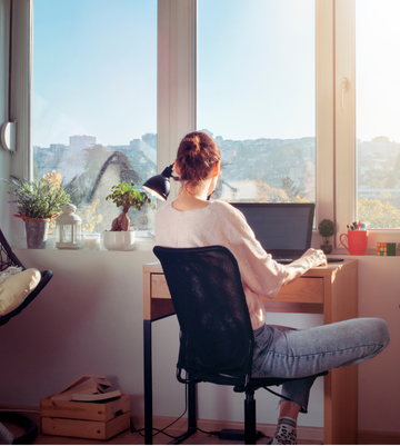Femme assise à un bureau près d'une grande fenêtre, travaillant sur un ordinateur portable avec une vue sur la ville.