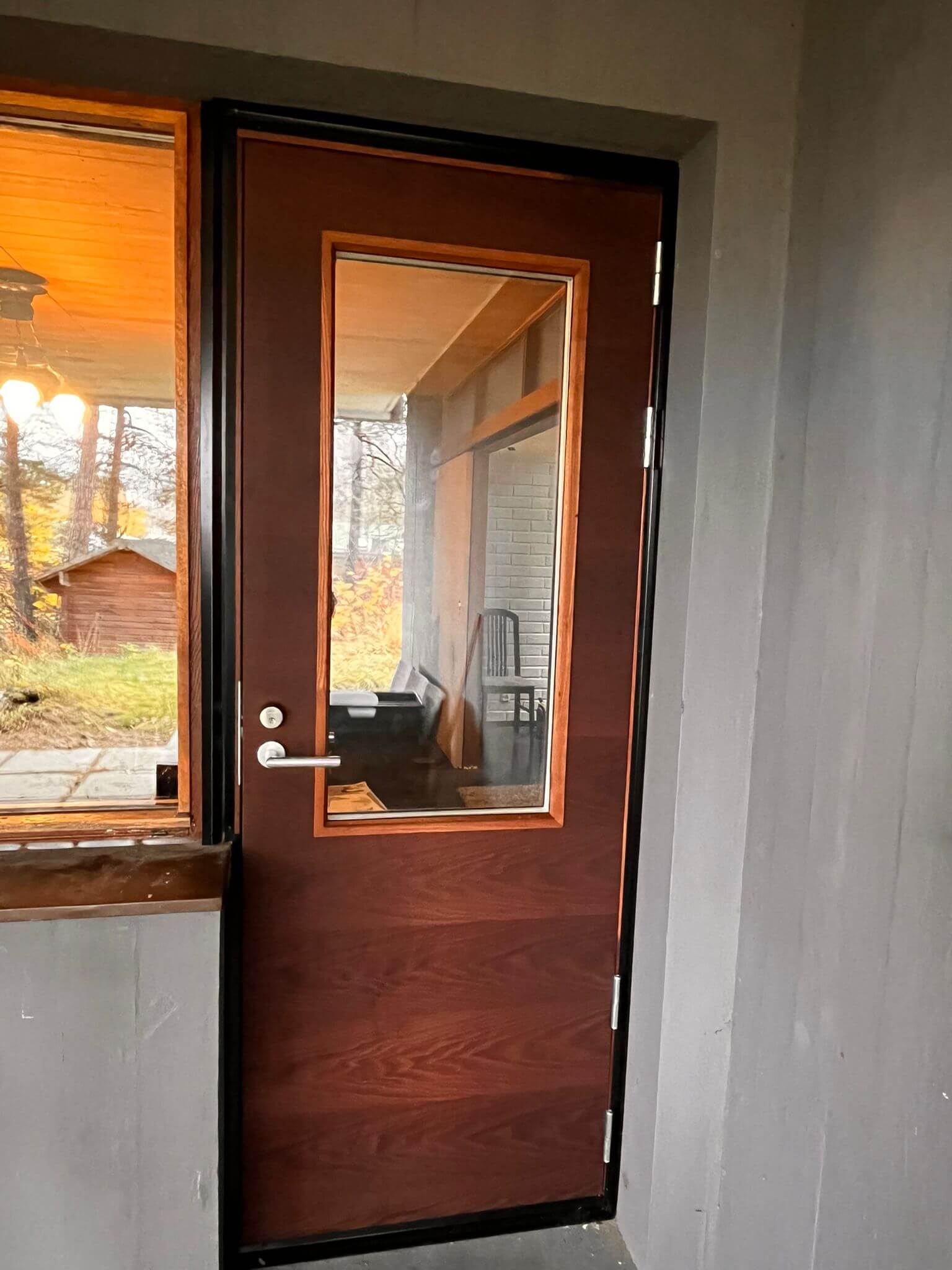 Tumma puinen ovi, jossa keskellä suuri suorakaiteen muotoinen ikkuna ja hopeanvärinen ovenkahva, ympäröivä harmaa seinä ja ikkunasta näkyy syksyinen metsämaisema.