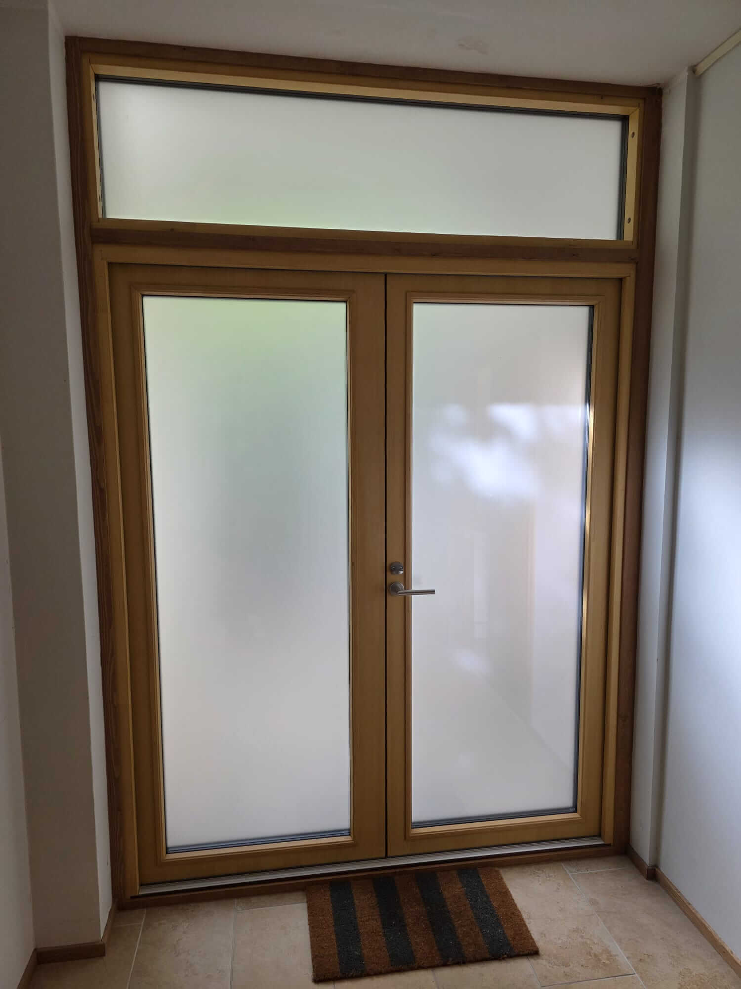 Kaksiosainen puuovi ja yläikkuna, joissa molemmissa on huurrettu lasi ja oven edessä matsiraita matto.