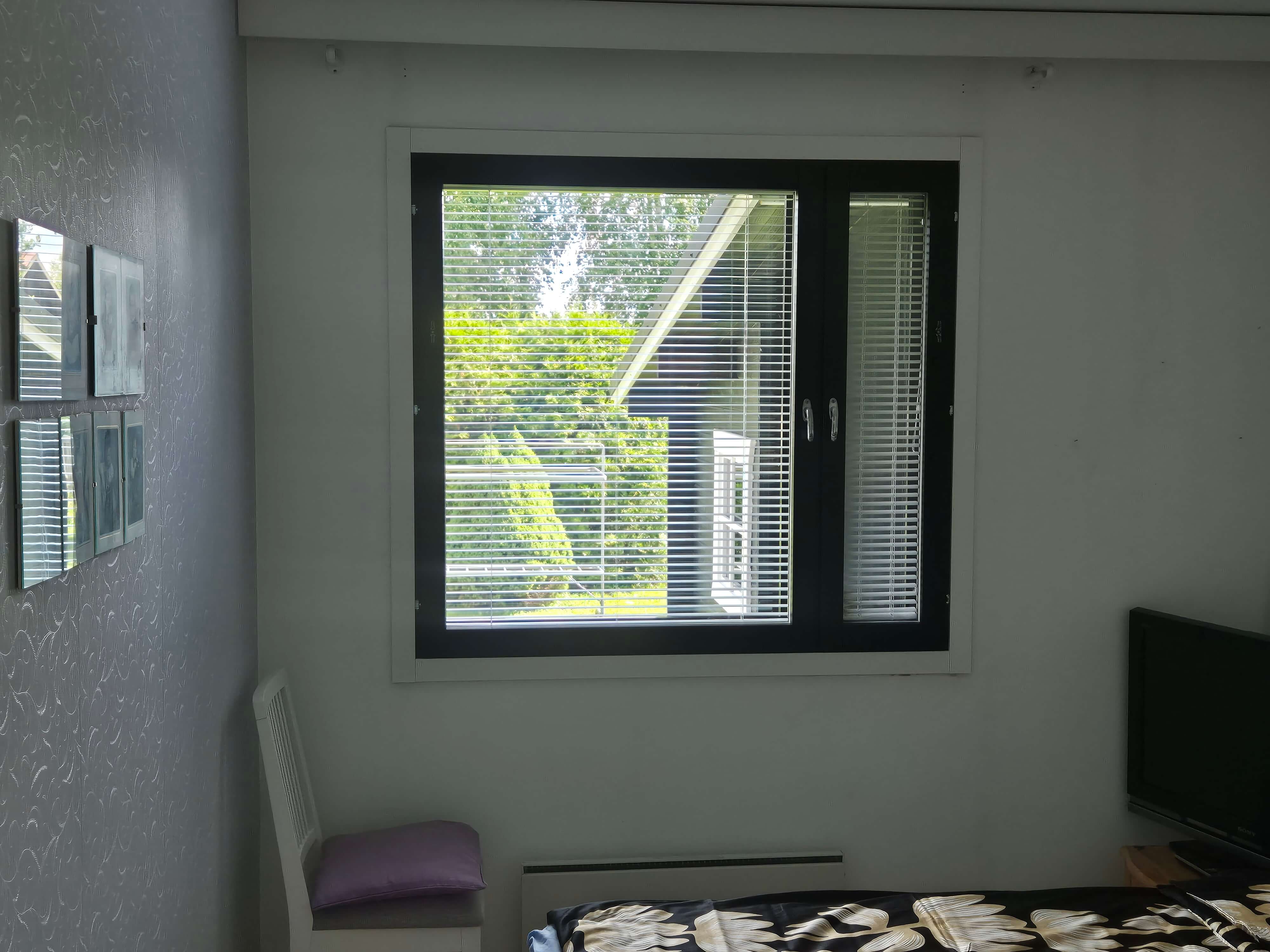 Huone, jossa seinässä ikkuna, ikkunassa valkoinen kaihdin, vasemmalla valkoinen tuoli, oikealla televisio ja alhaalla osa vuoteesta mustalla ja keltaisella kukkakuvioidulla peitolla.