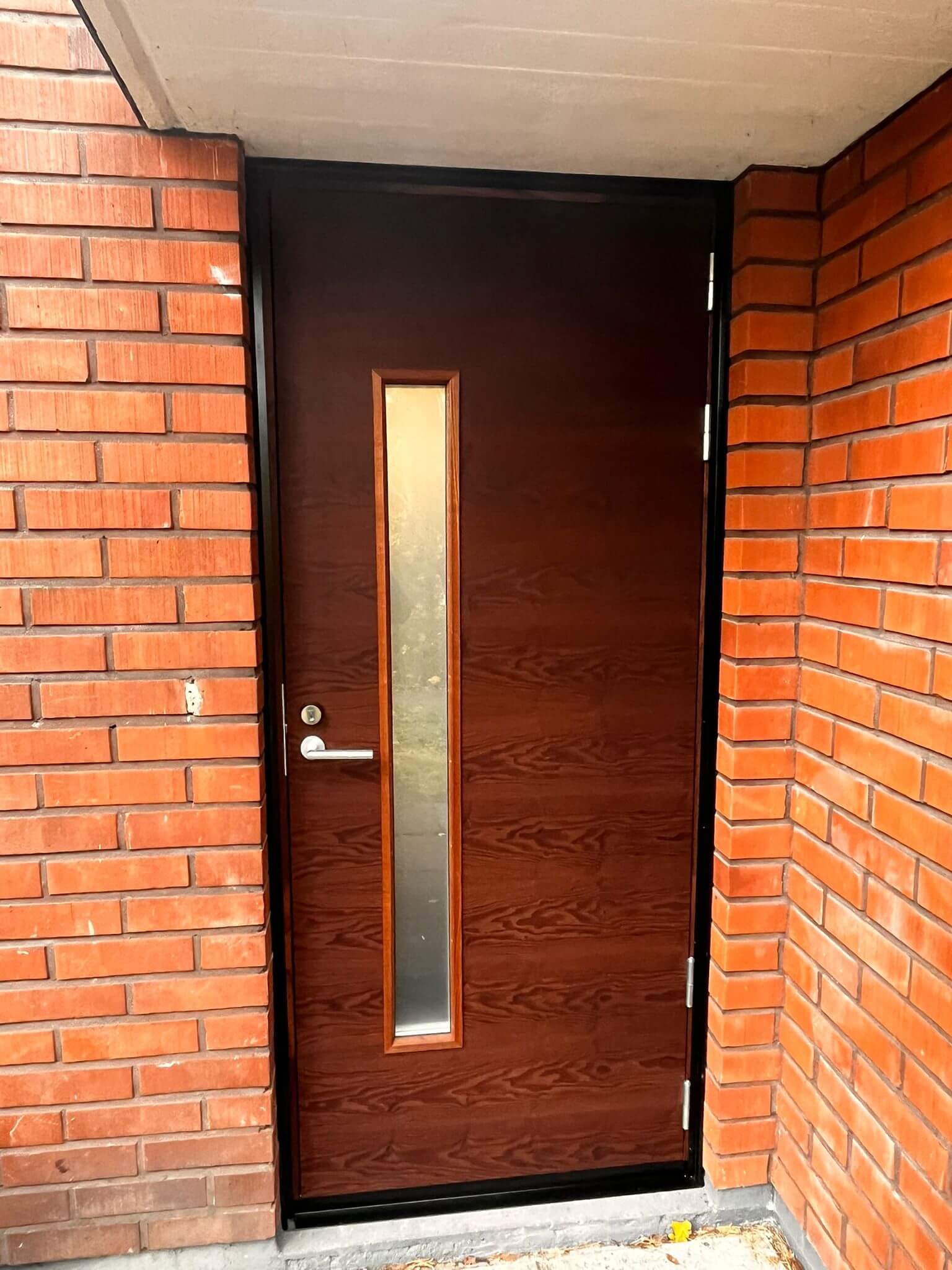 Tumma puuovi, jossa kapea pystysuora ikkuna ja metallinen ovenkahva, oranssien tiiliseinien välissä.