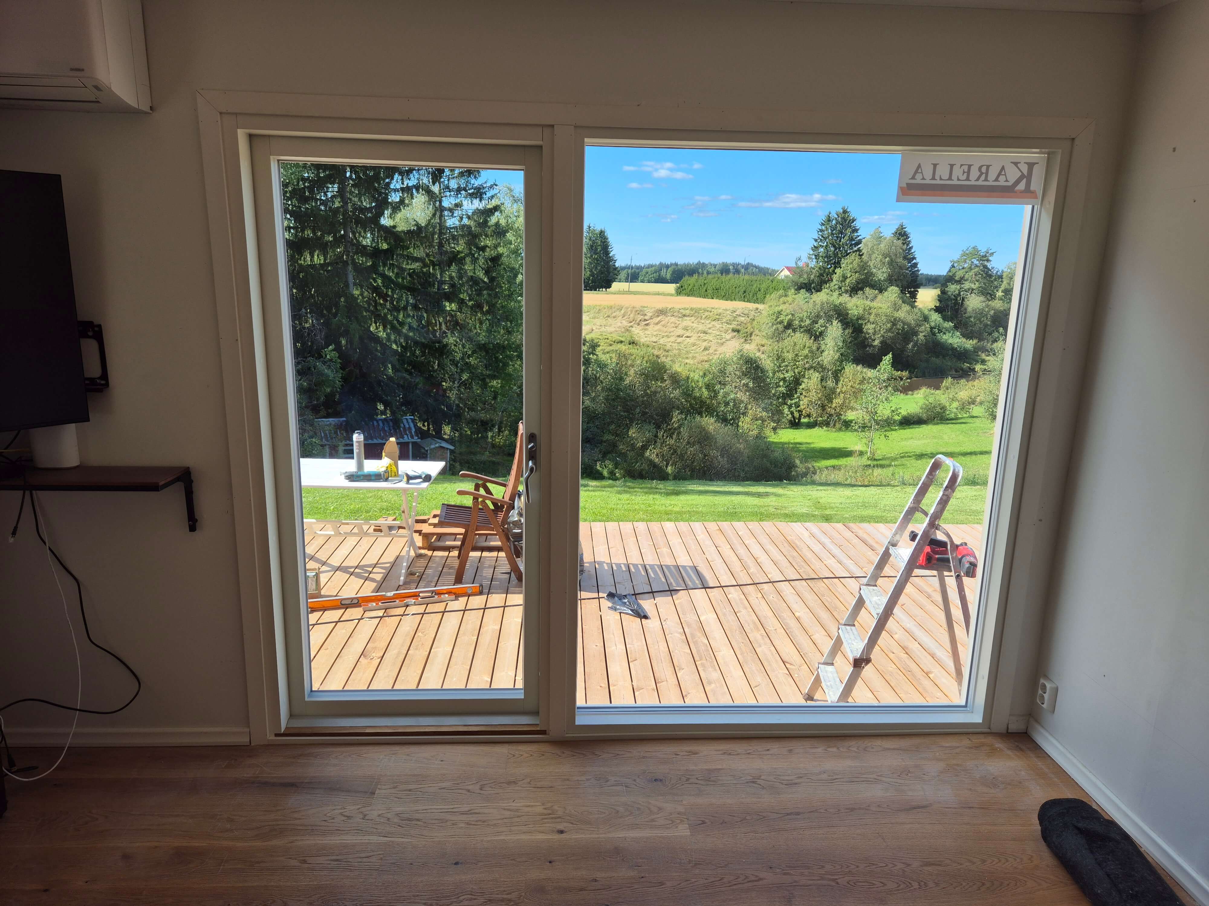 Näkymä lasiovesta, joka avautuu puiselle terassille, jossa on tikapuut ja puutarhatuolit, taustalla vihreä niitty ja puita.