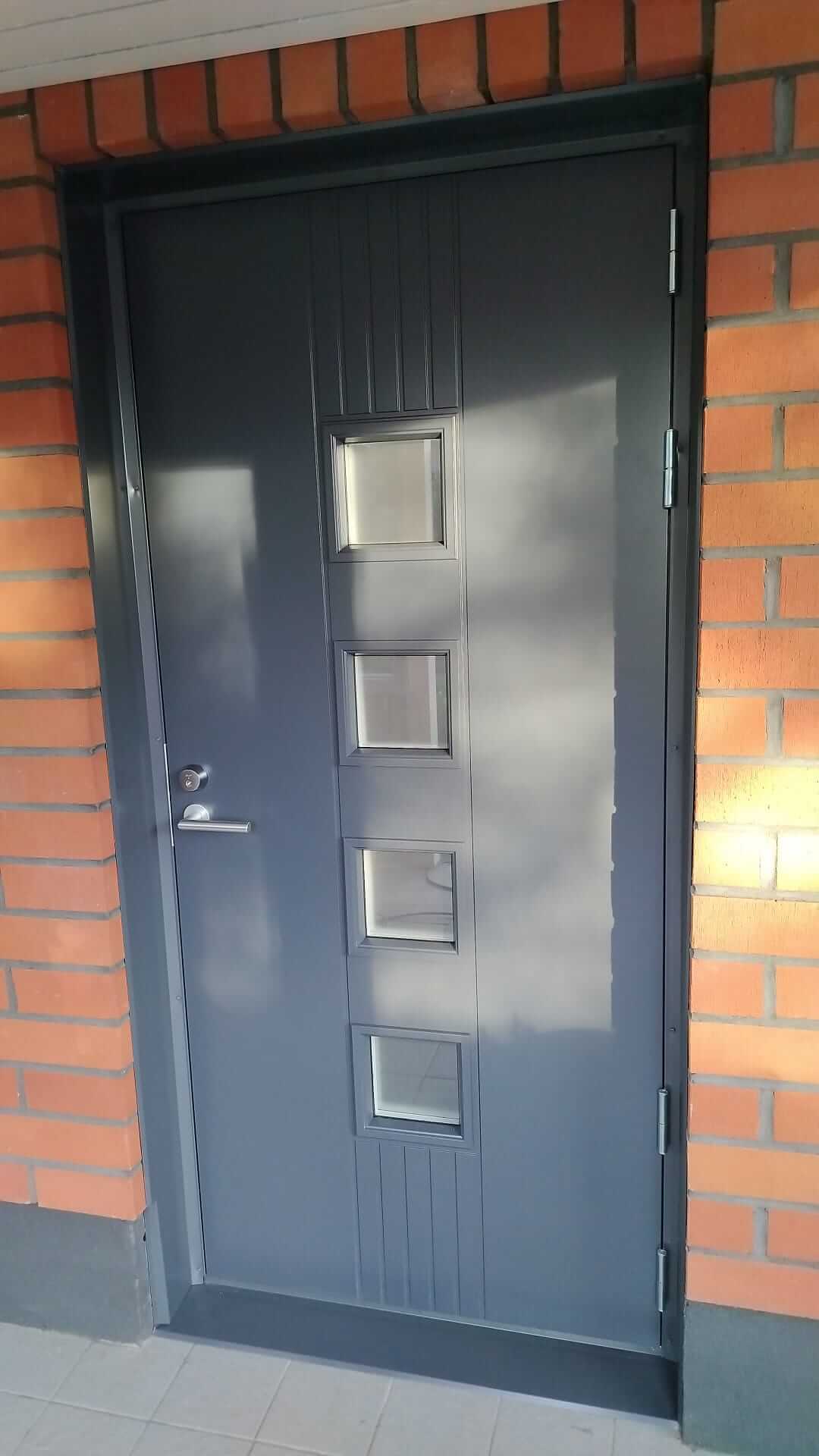 Tummanharmaa ulko-ovi, jossa neljä pientä neliönmuotoista ikkunaa pystysuorassa rivissä, ympärillä tiiliseinää.