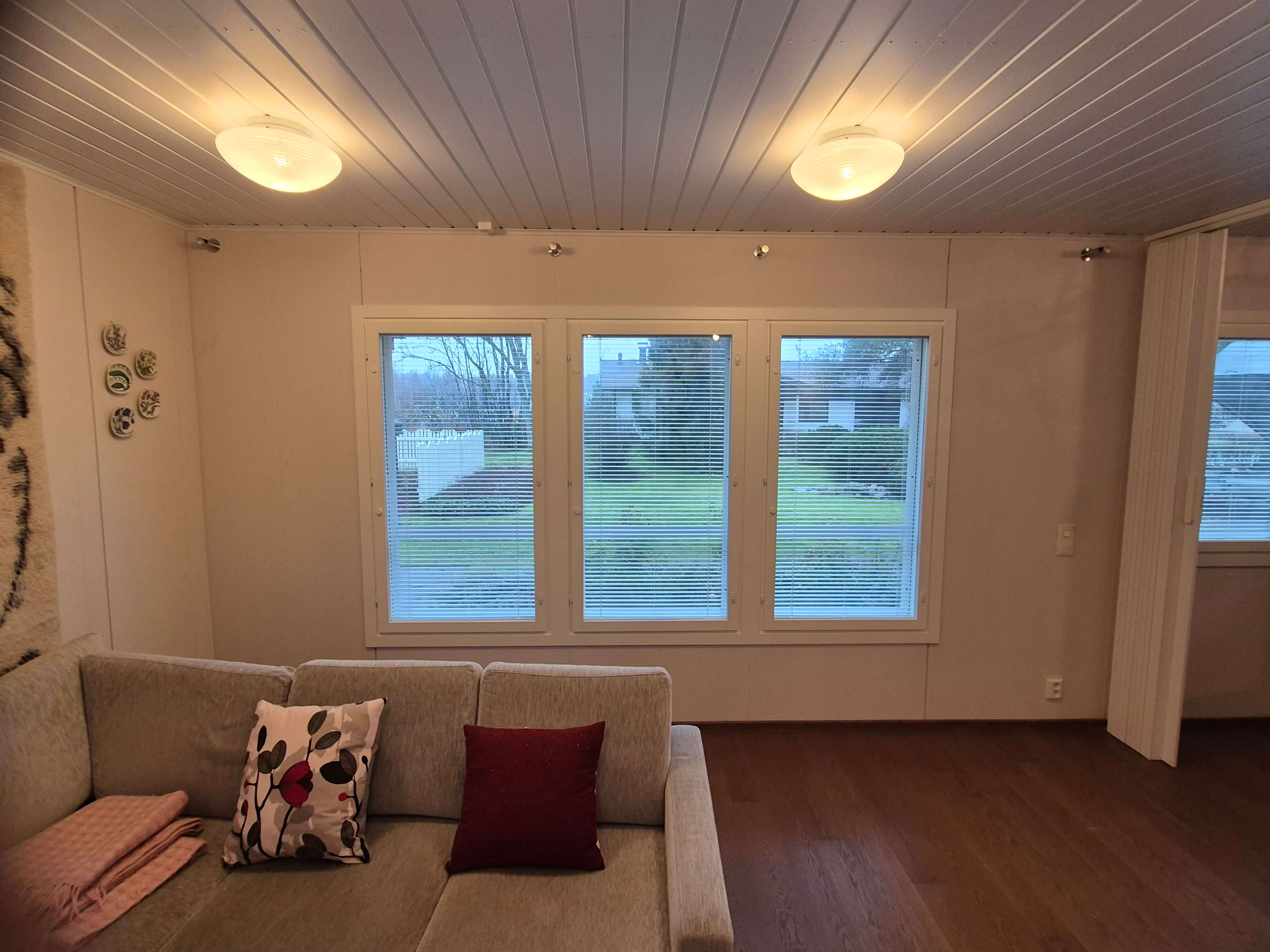 Vaaleanharmaa kulmasohva, jossa kaksi koristetyynyä ja vaaleanpunainen viltti, ikkunaseinä, joka avautuu näkymään nurmikentälle ja taloihin.