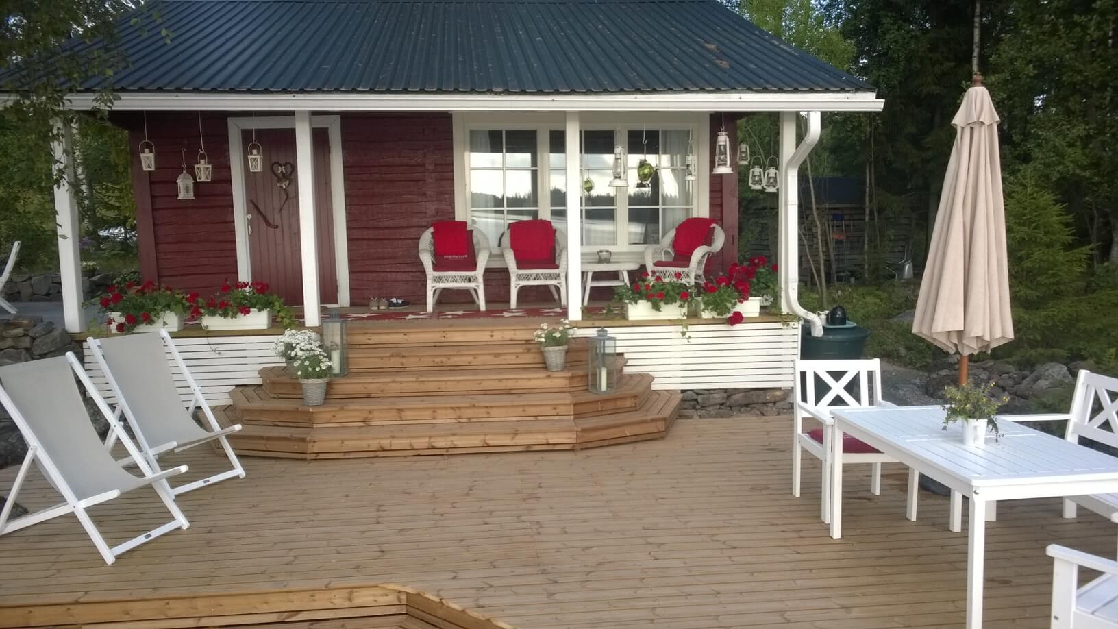 Puinen terassi, jossa kolme valkoista nojatuolia punaisilla tyynyillä ja kukkia rapuissa punaisen mökin edessä.