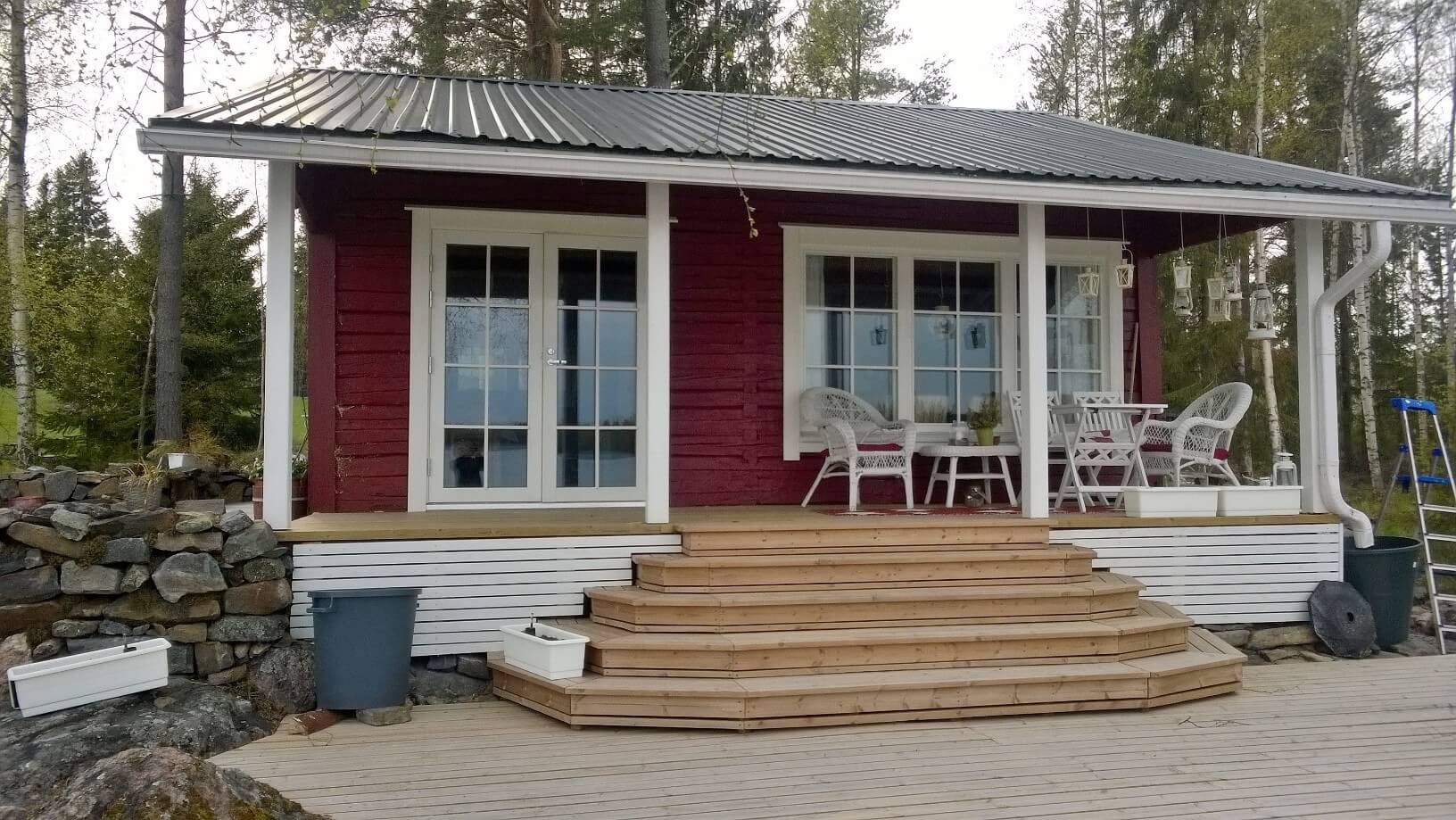 Punainen mökki valkoisin ikkuna- ja ovikehyksin, puiset portaat ja terassi, valkoisia puutarhatuoleja ja pöytä, taustalla metsä.