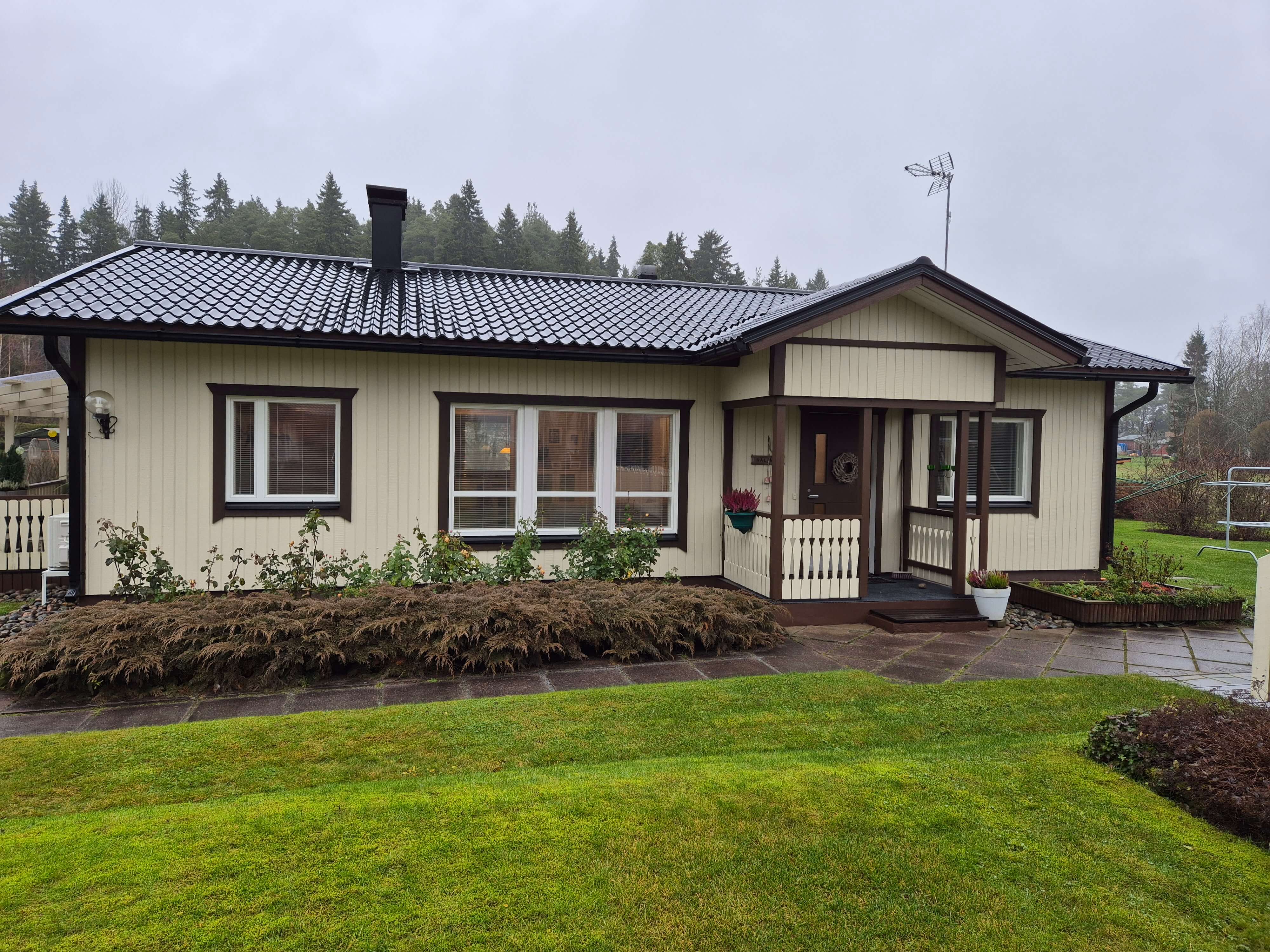Yksikerroksinen vaalea puutalo tumman katon ja pienen kuistin kanssa, edessä nurmikko ja pensasaita.