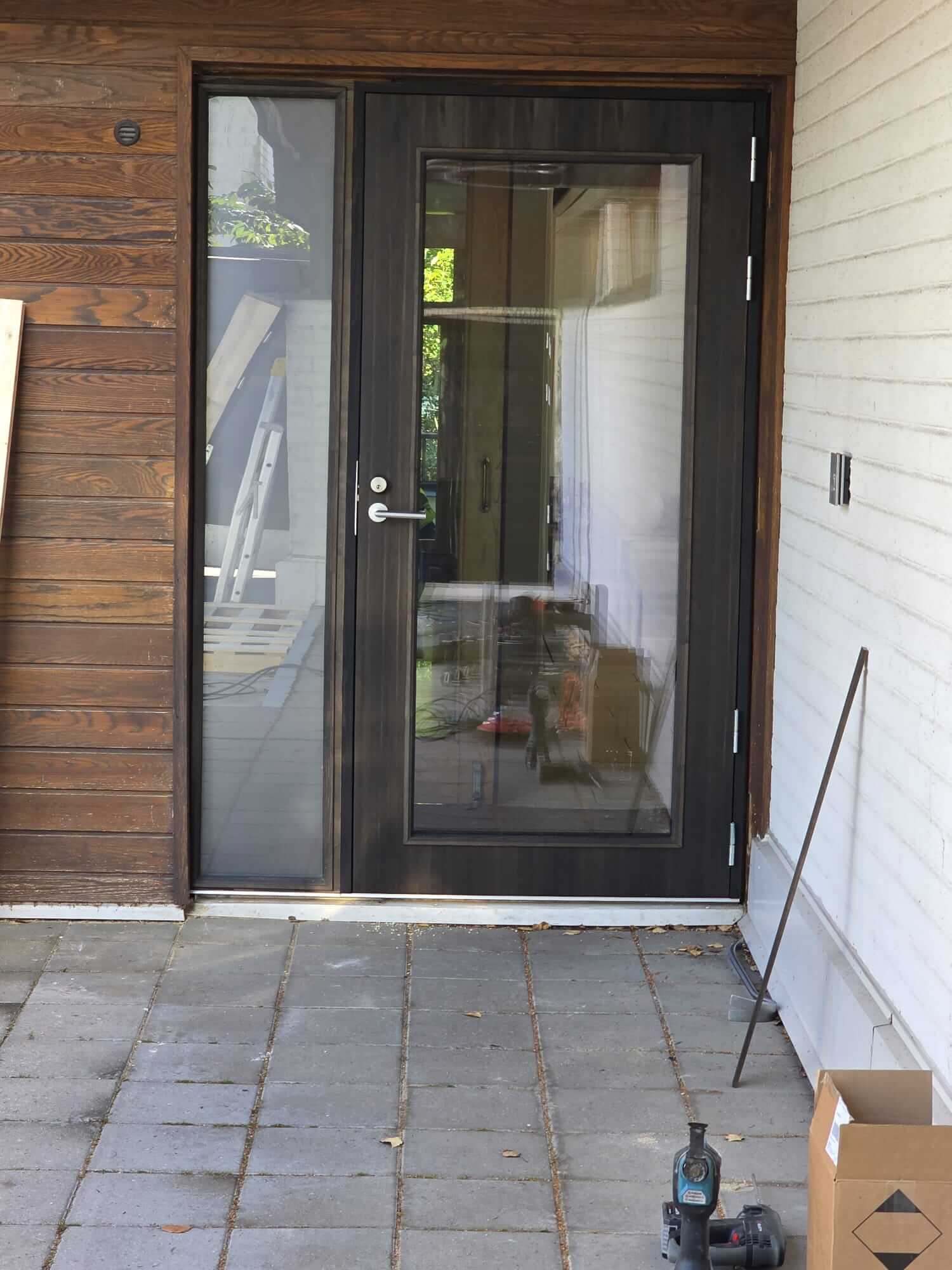 Musta lasiovi, jossa vasemmalla kapea lasiikkuna, edessä harmaata laattalattiaa ja oikealla sähkötyökalu ja pahvilaatikko.