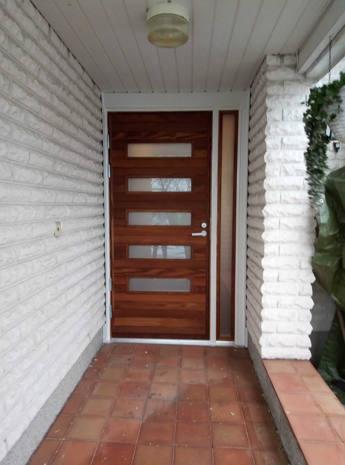 Sisäänkäynti, jossa on ruskea puuovi, jossa viisi vaakasuuntaista lasiruudua, valkoinen tiiliseinä ja terrakottanruskea laattalattia.