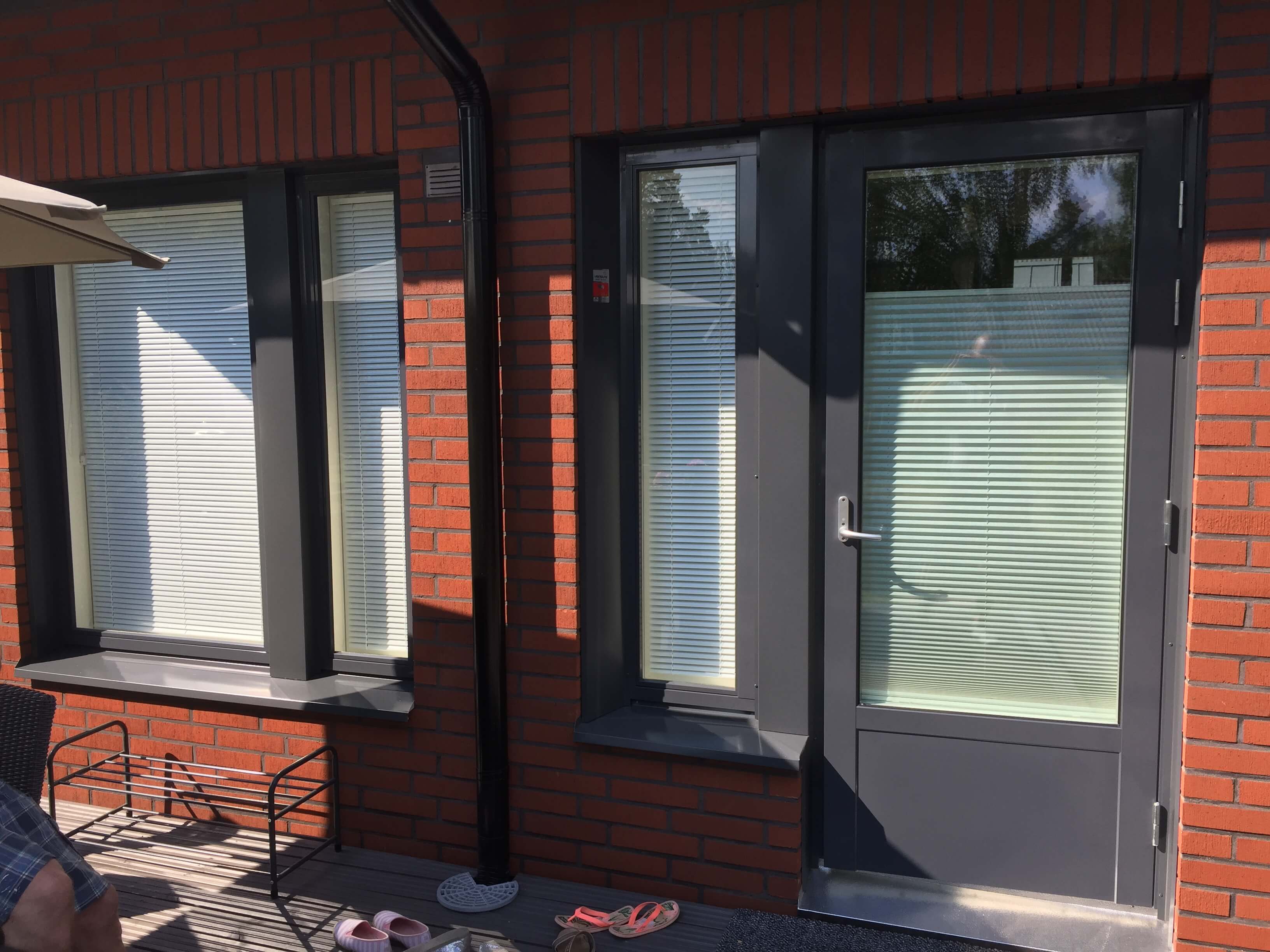 Ulkopatio, jossa punatiilinen talon julkisivu, harmaa ovi ja kaksi ikkunaa, sekä tossut ja metallinen kenkäteline.