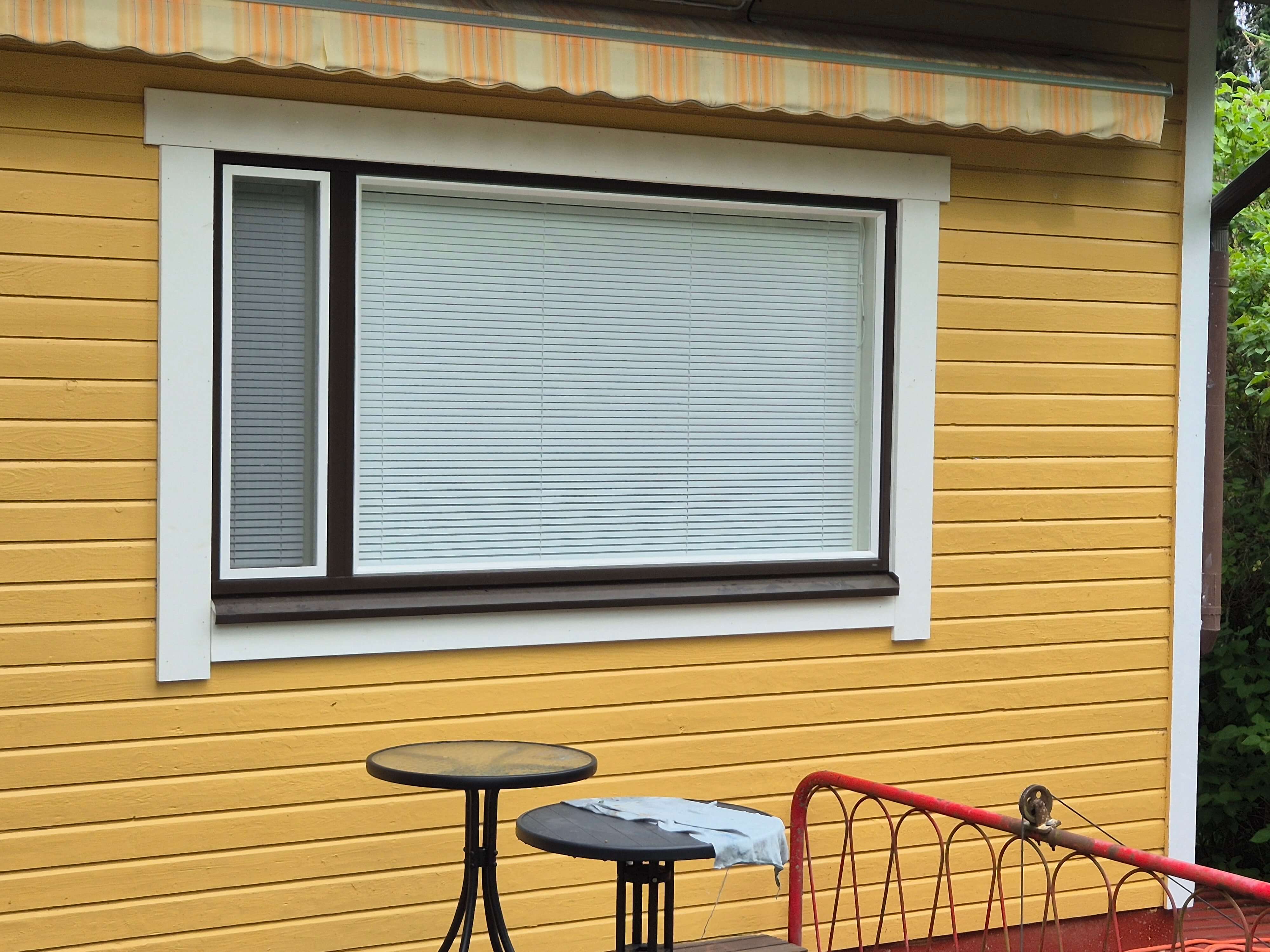 Keltaisen puutalon seinä, jossa ikkuna ja verho, edessä kaksi mustaa pyöreää ulkopöytää ja punainen metallikaide.