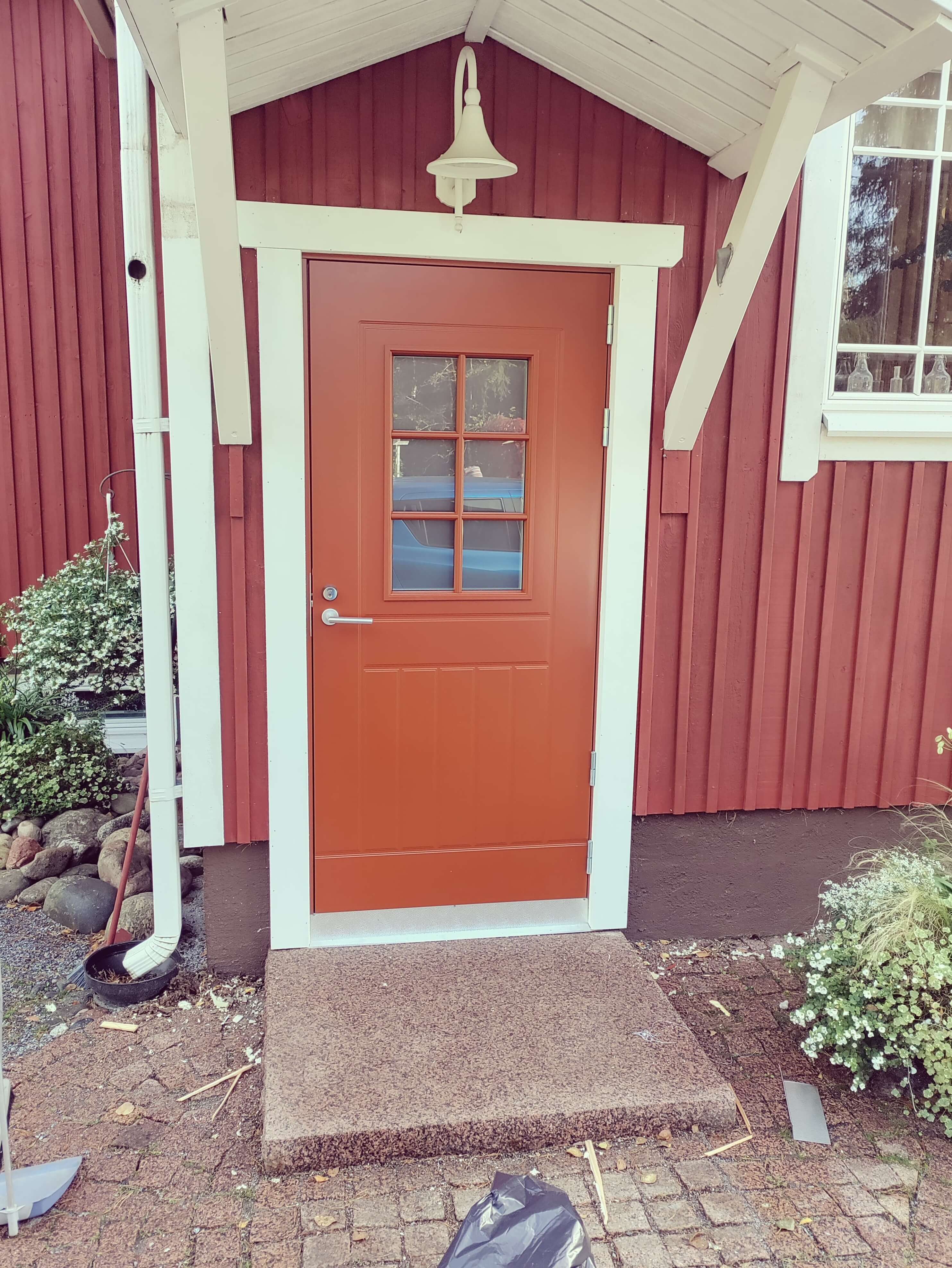 Punainen ulko-ovi, jossa on pieni ruudullinen ikkuna, valkoiset kehykset ja valkoinen ulkovalo talon punaisessa laudassa.