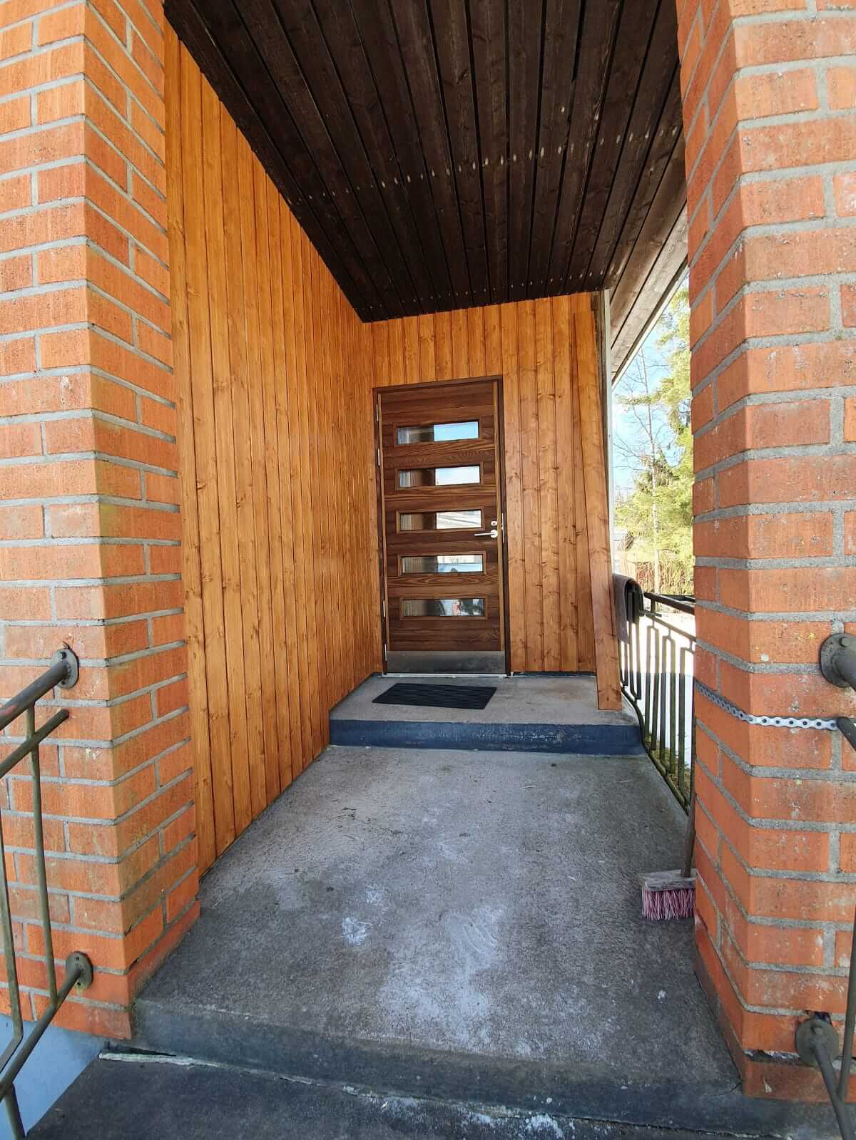 Sisäänkäynti puuseinillä ja -ovella, tiiliset pylväät ja betoninen lattia porrasaskelmineen.