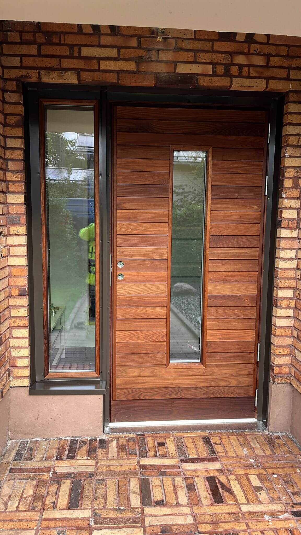 Etupuoli, jossa moderni puinen ovi pystysuuntaisella kapealla ikkunalla ja sen vasemmalla puolella kapea ikkunaruutu, tiiliseinän kehystämänä.