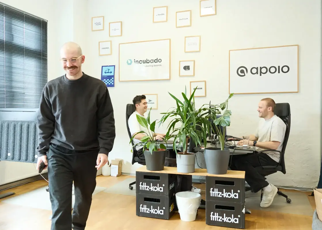 2 men sitting at desk and 1 man walking towards camera