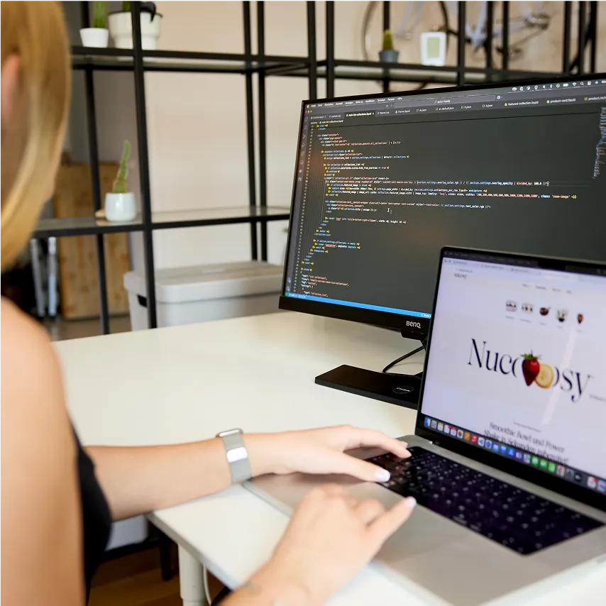 women sitting at office desk looking at code on monitor and laptop