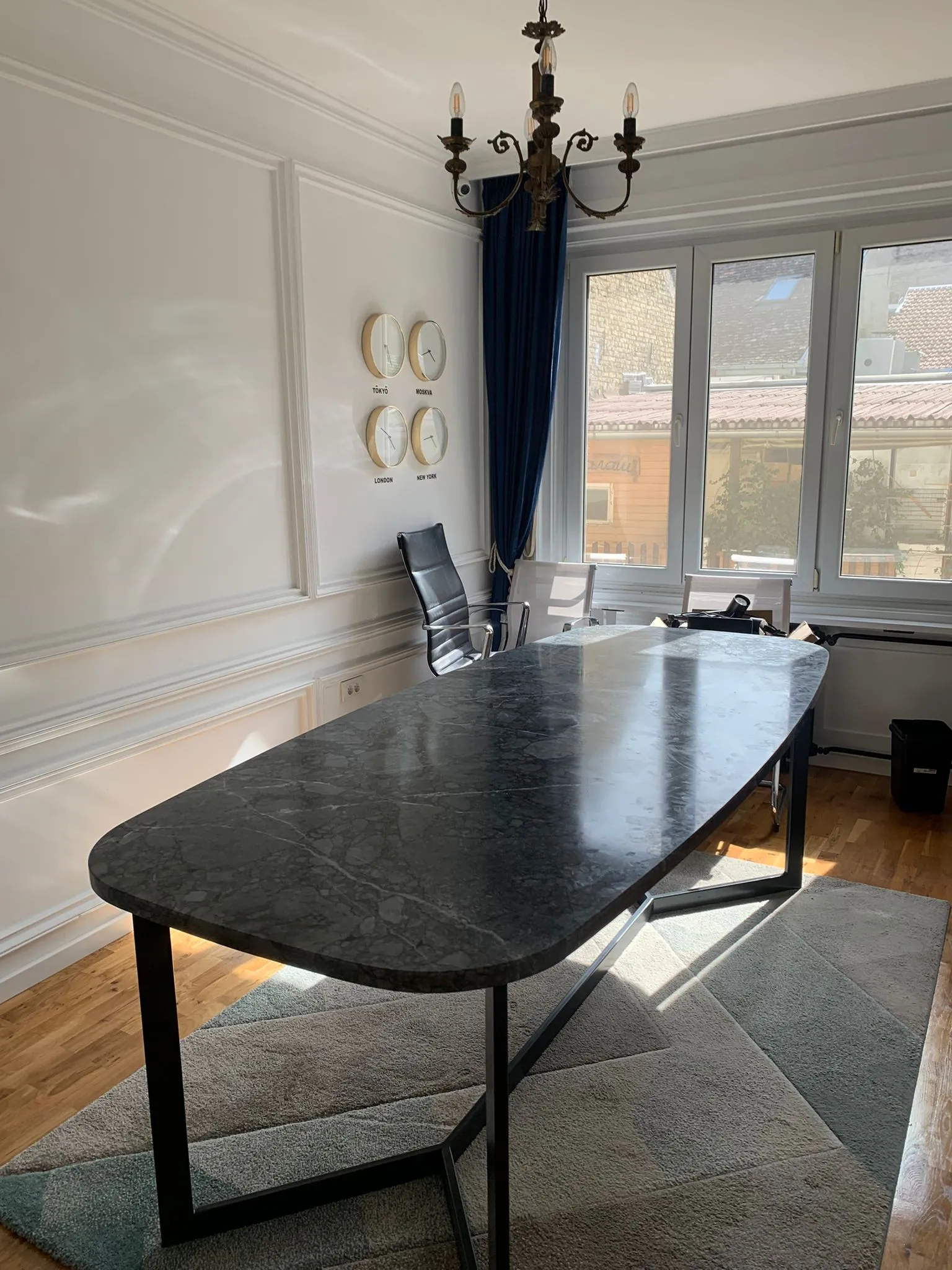 Daqaaqaan xafiiska casriga ah oo leh miis buluug madow ah, kursi madow oo raaxo leh, saacadyo darbiga ku dheggan oo muujinaya saacadaha magaalooyinka Tokyo, Moscow, London, iyo New York, iyo daaqado waaweyn oo leh daahyada buluugga madow.