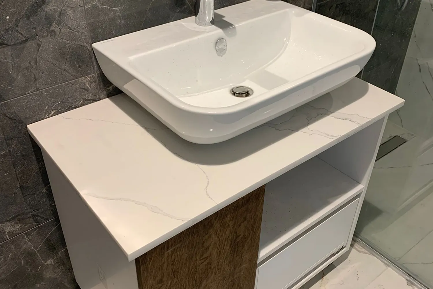 White rectangular bathroom sink mounted on a countertop with light marble pattern and a wooden cabinet below.