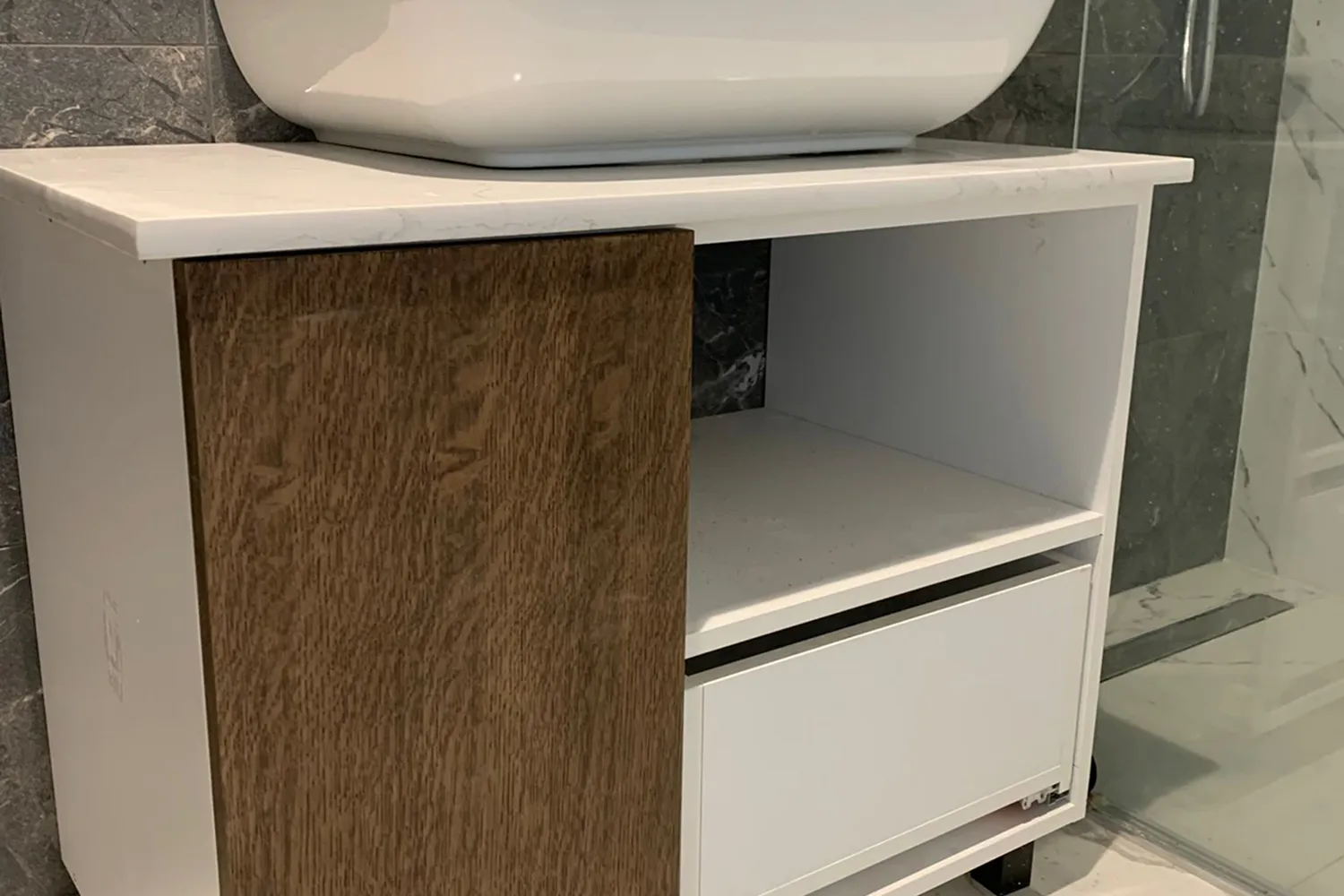 White bathroom vanity with a wood grain door, open shelf, and drawer next to a glass shower enclosure.