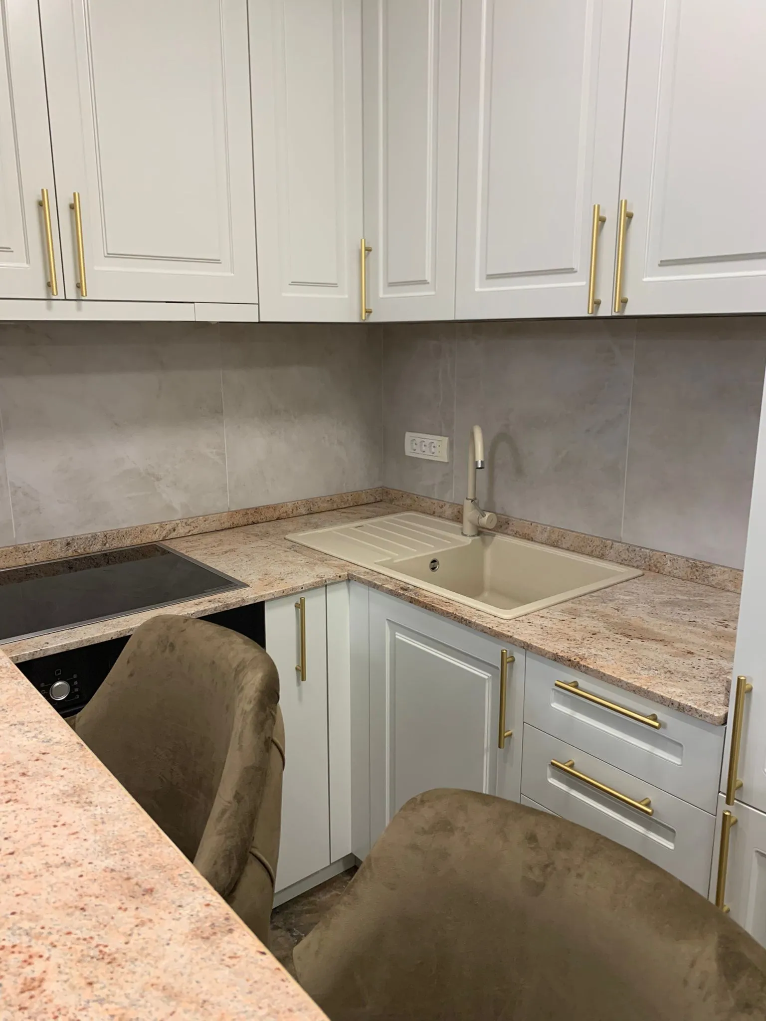 Kitchen corner with beige countertop, white cabinets with gold handles, a beige sink, and two brown upholstered chairs.