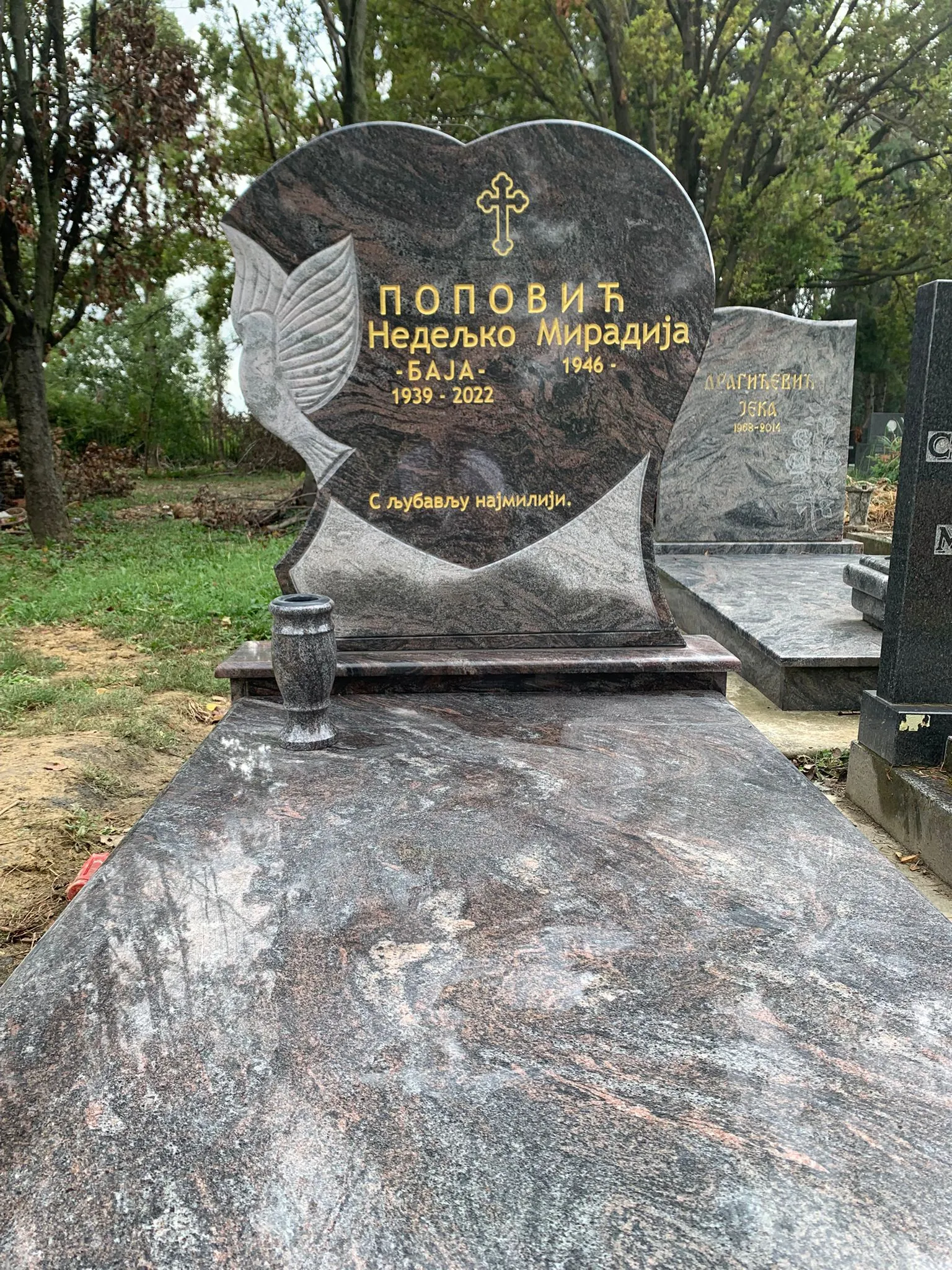 Grave with a heart-shaped headstone engraved with a cross, a dove, and Serbian text marking the name Nedeljko Miradija Popović with years 1939-2022, set in a forested cemetery.