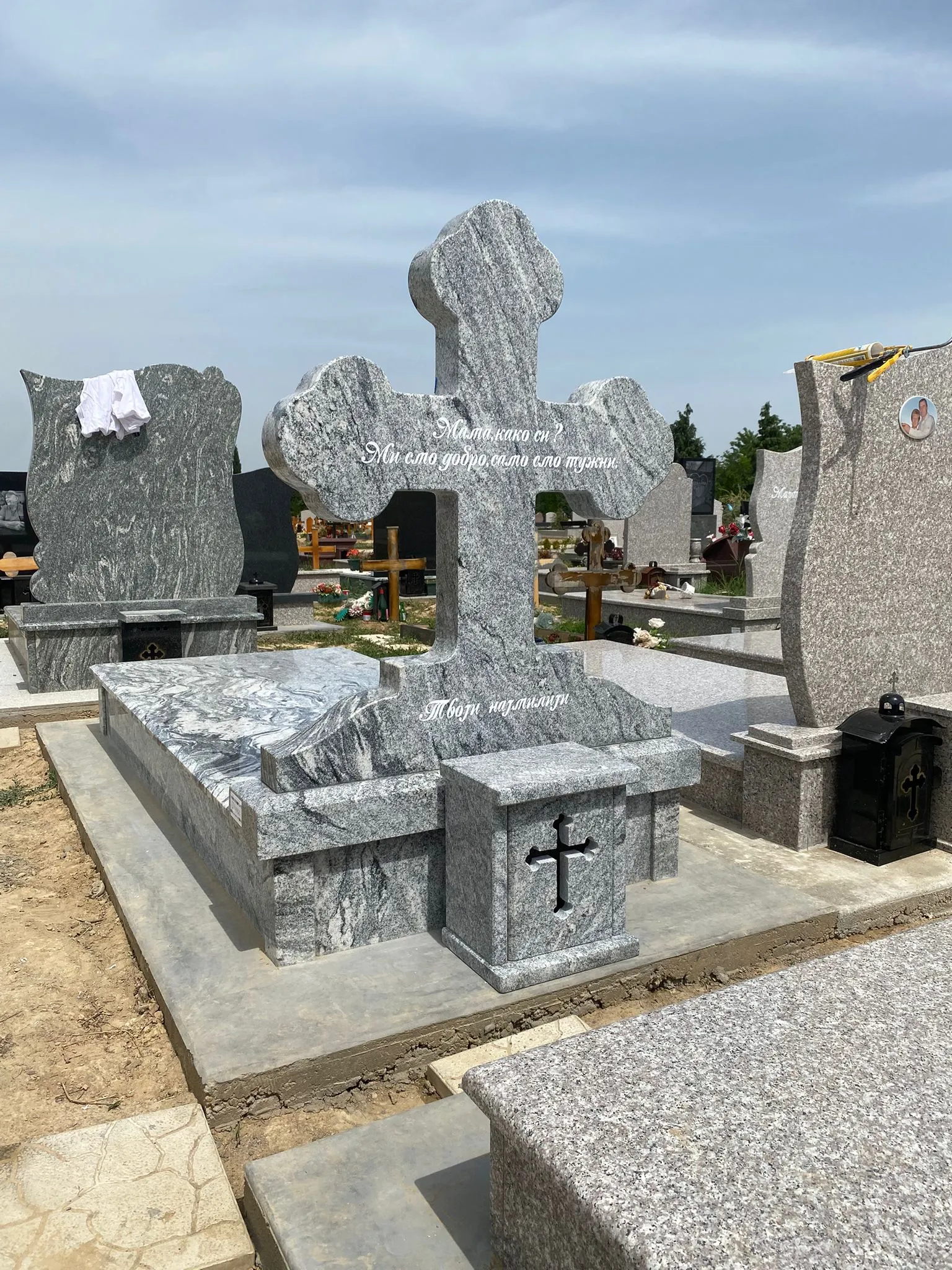 Grave with a large gray stone cross and engraved text, surrounded by other gravestones in a cemetery under a partly cloudy sky.