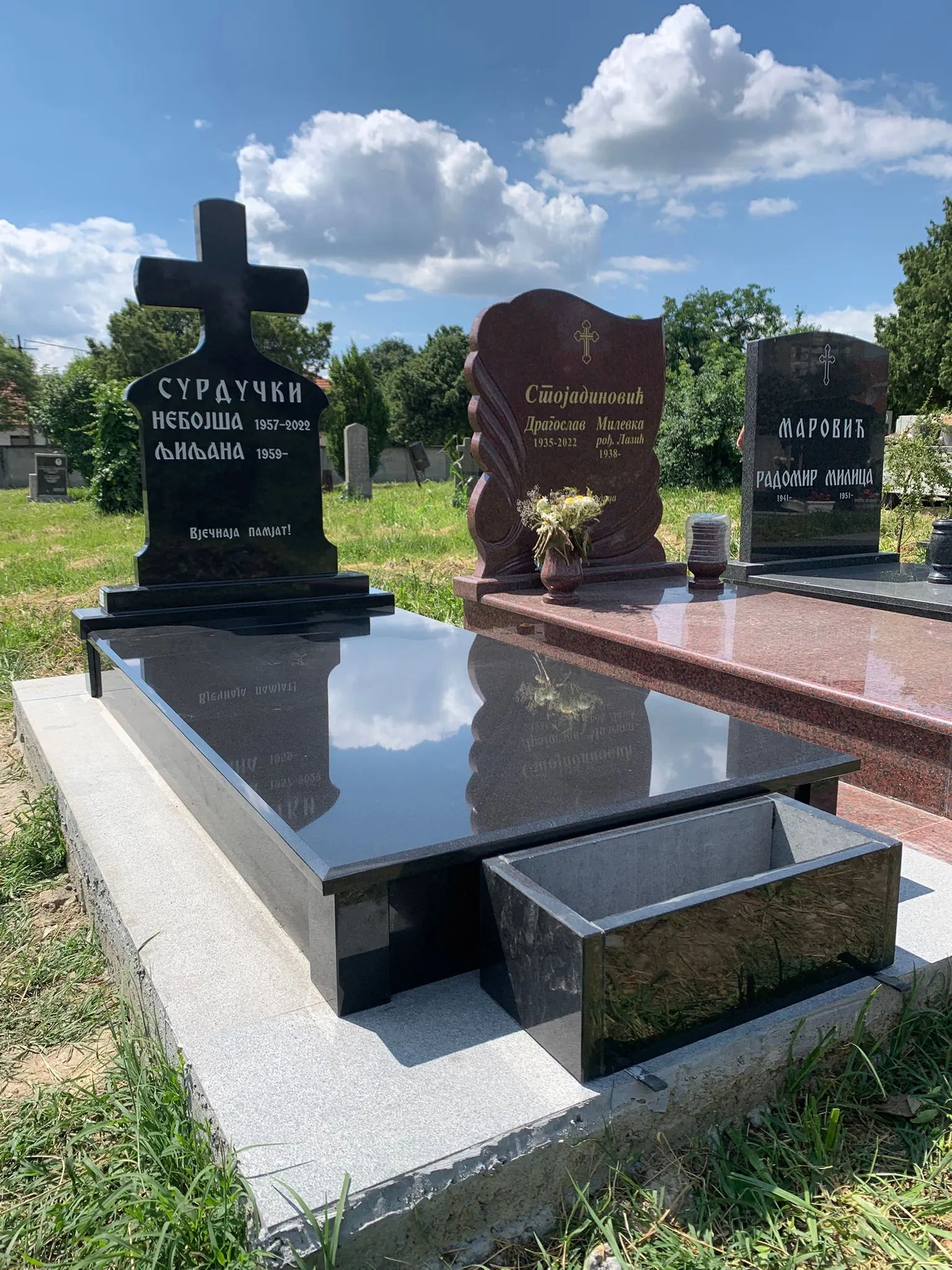Three polished headstones in a grassy cemetery under a partly cloudy sky, with one black grave reflecting clouds and adjacent dark flower vase.