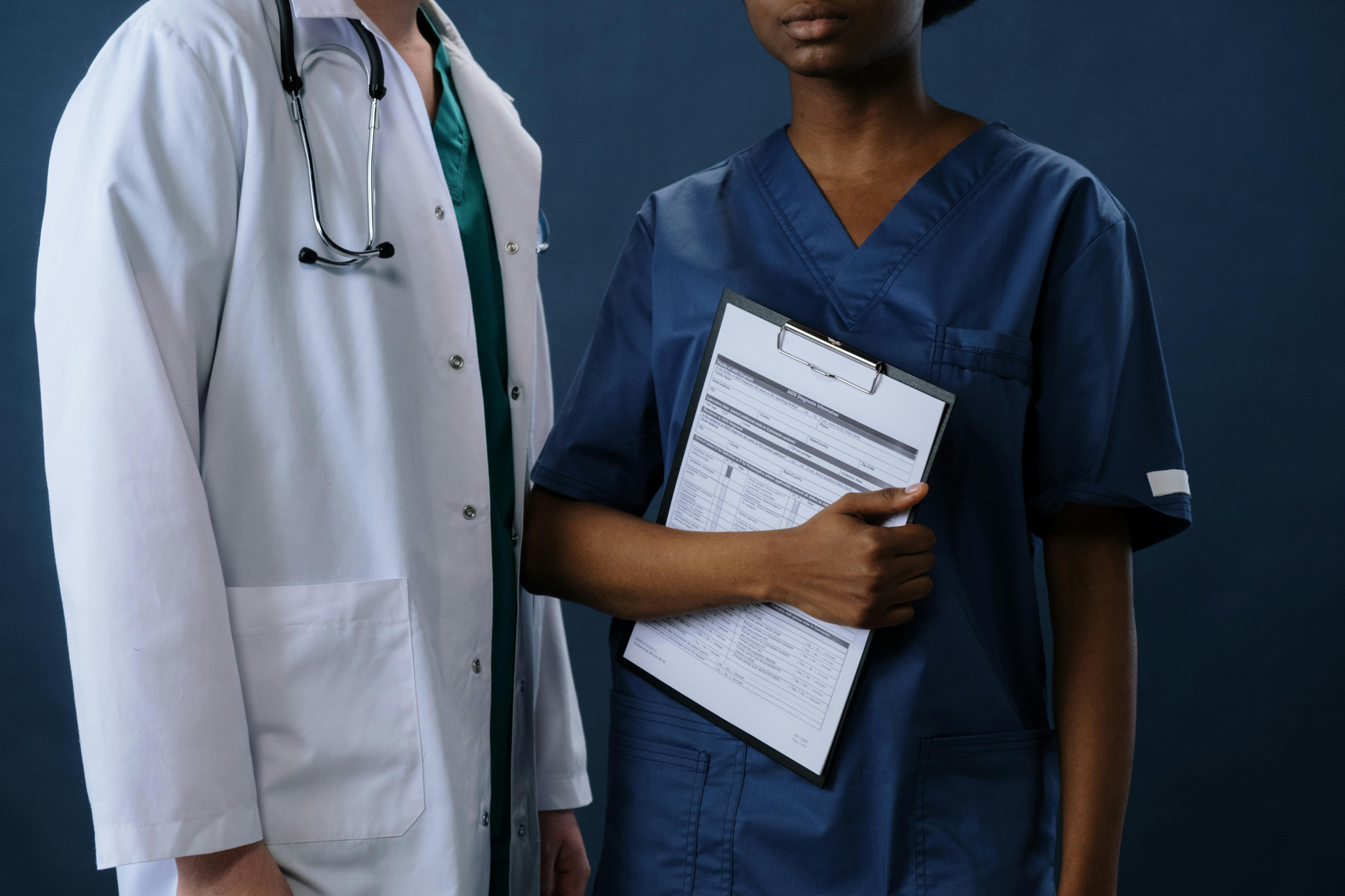 two faceless people, person on the left side has a doctor coat and a stethoscope. the person on the right is wearing blue scrubs and holding a clipboard