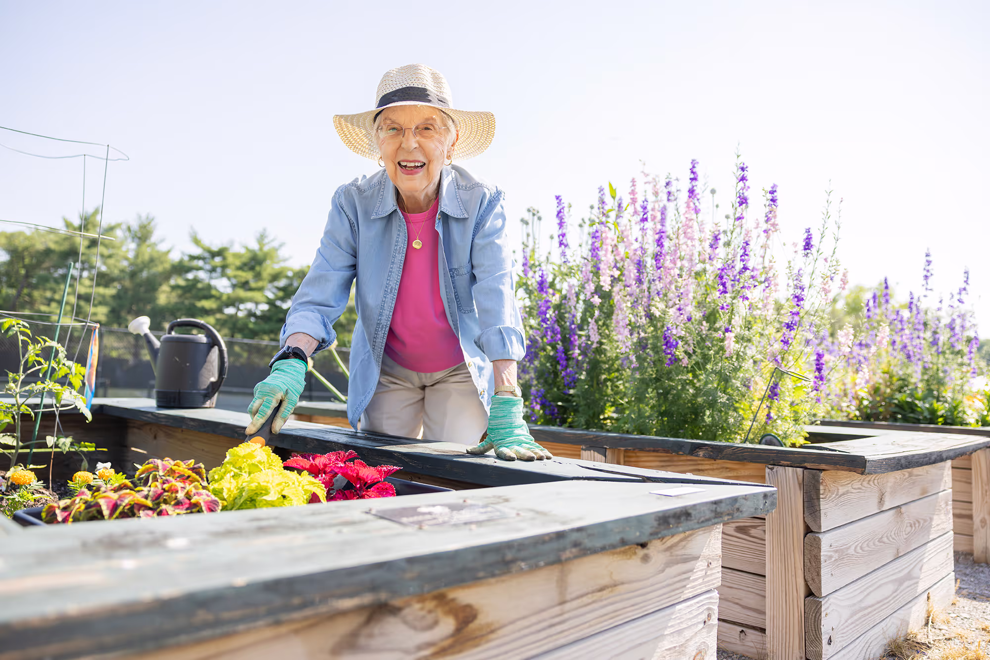 An elderly woman smiles while gardening in a raised bed, surrounded by flowers.