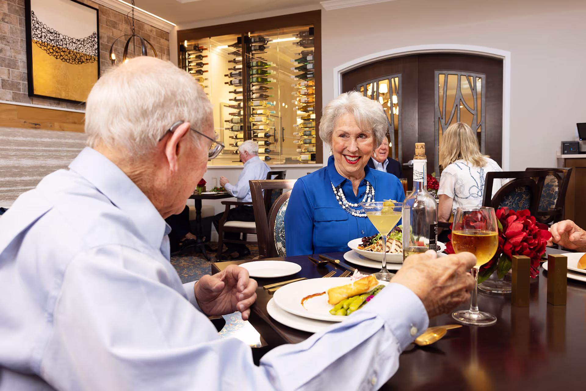 An older couple dines together at a restaurant, smiling and holding drinks.
