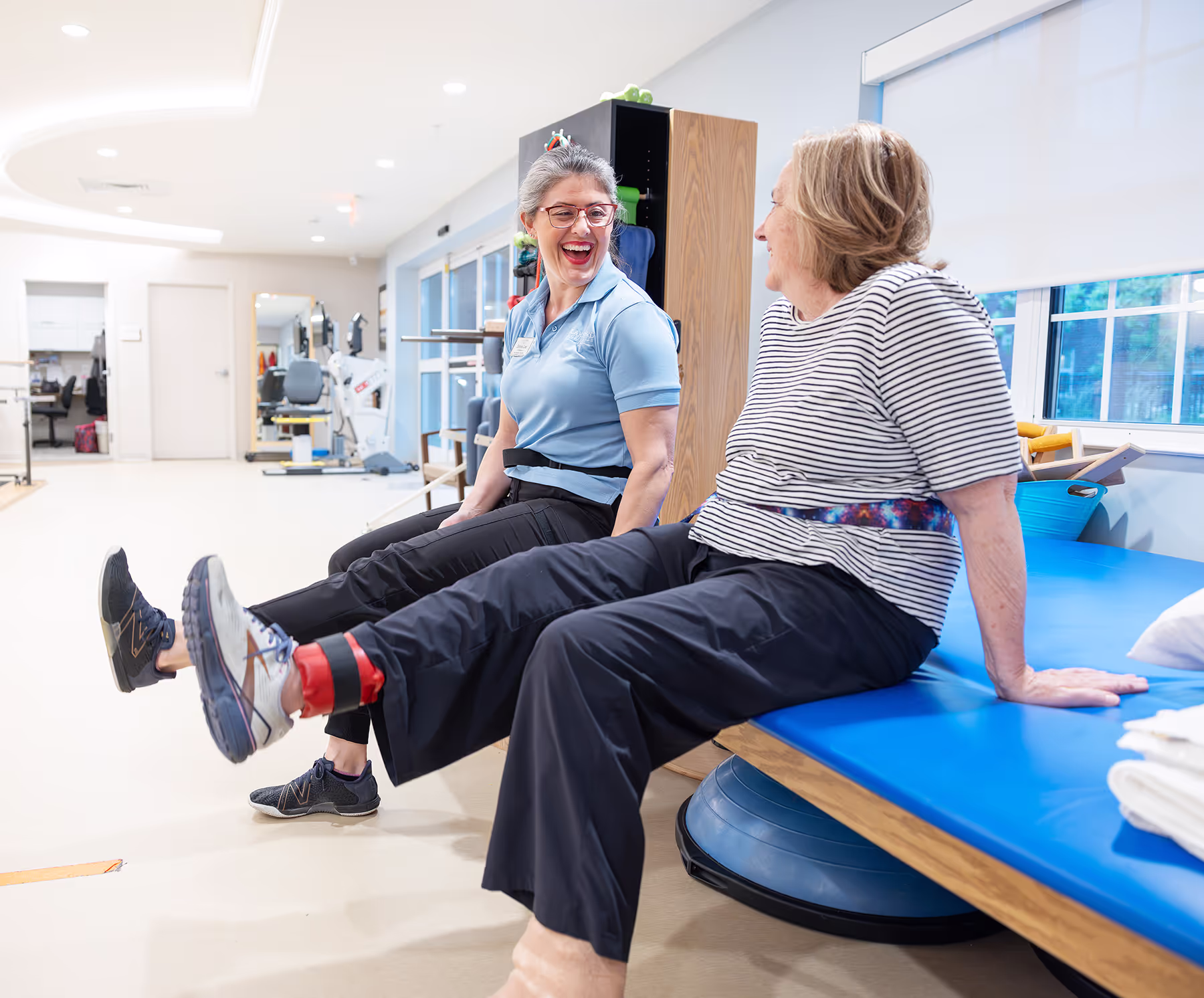 Two women in a gym, smiling and exercising on a blue platform.